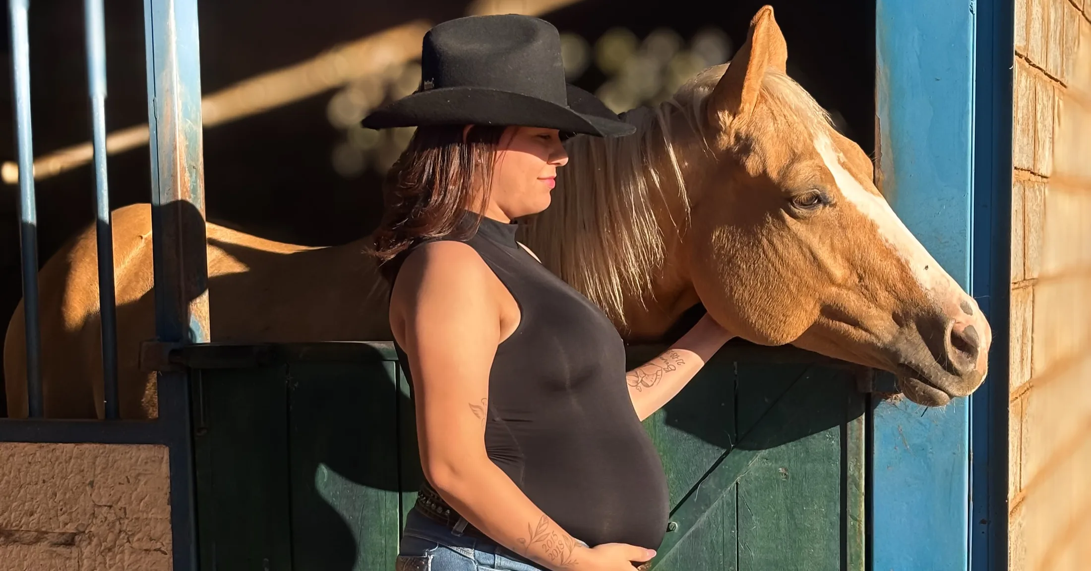Mulher de chapéu preto encosta a cabeça junto a um cavalo diante de uma estrutura de madeira.