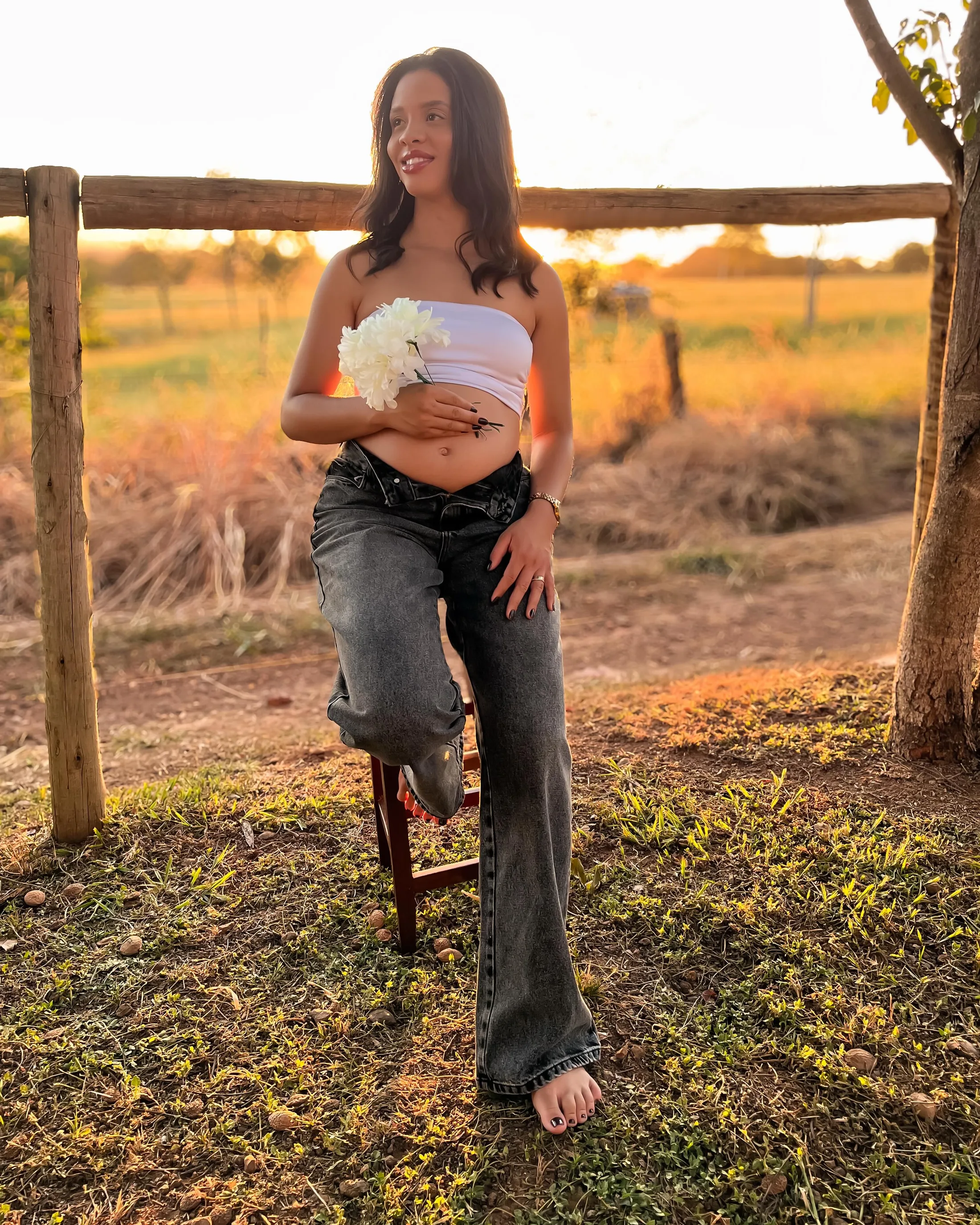 Mulher de top branco e calça jeans segura flores junto a uma cerca no entardecer.
