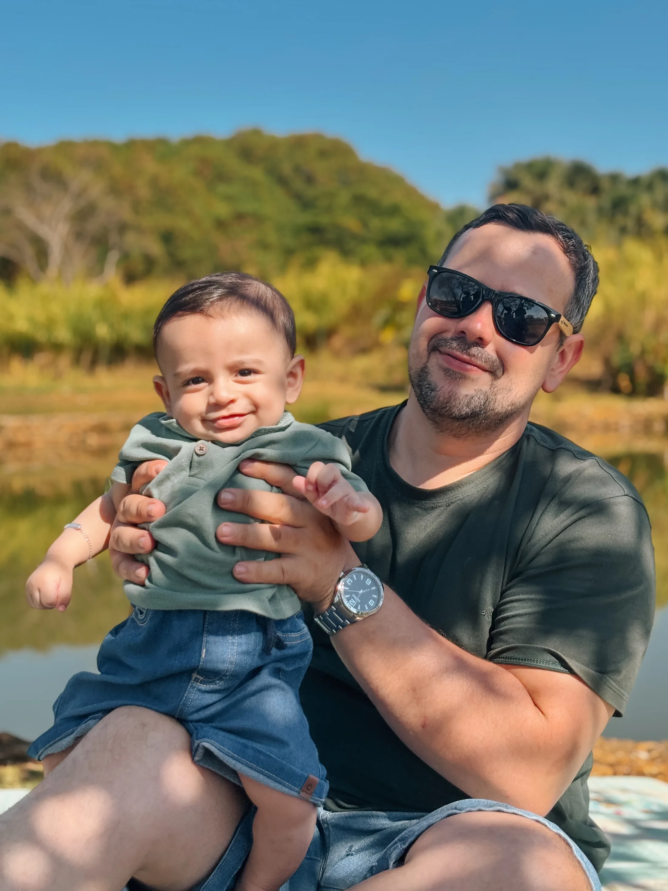 Homem de óculos escuros ergue um bebê de roupa verde em um campo ensolarado.