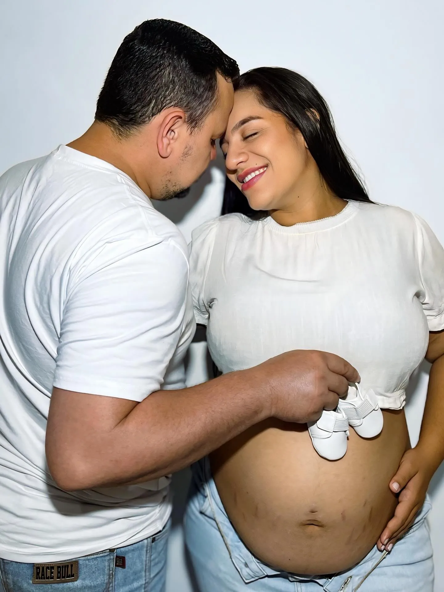 Casal de camiseta branca aproxima o rosto durante um ensaio de gestação em fundo claro.