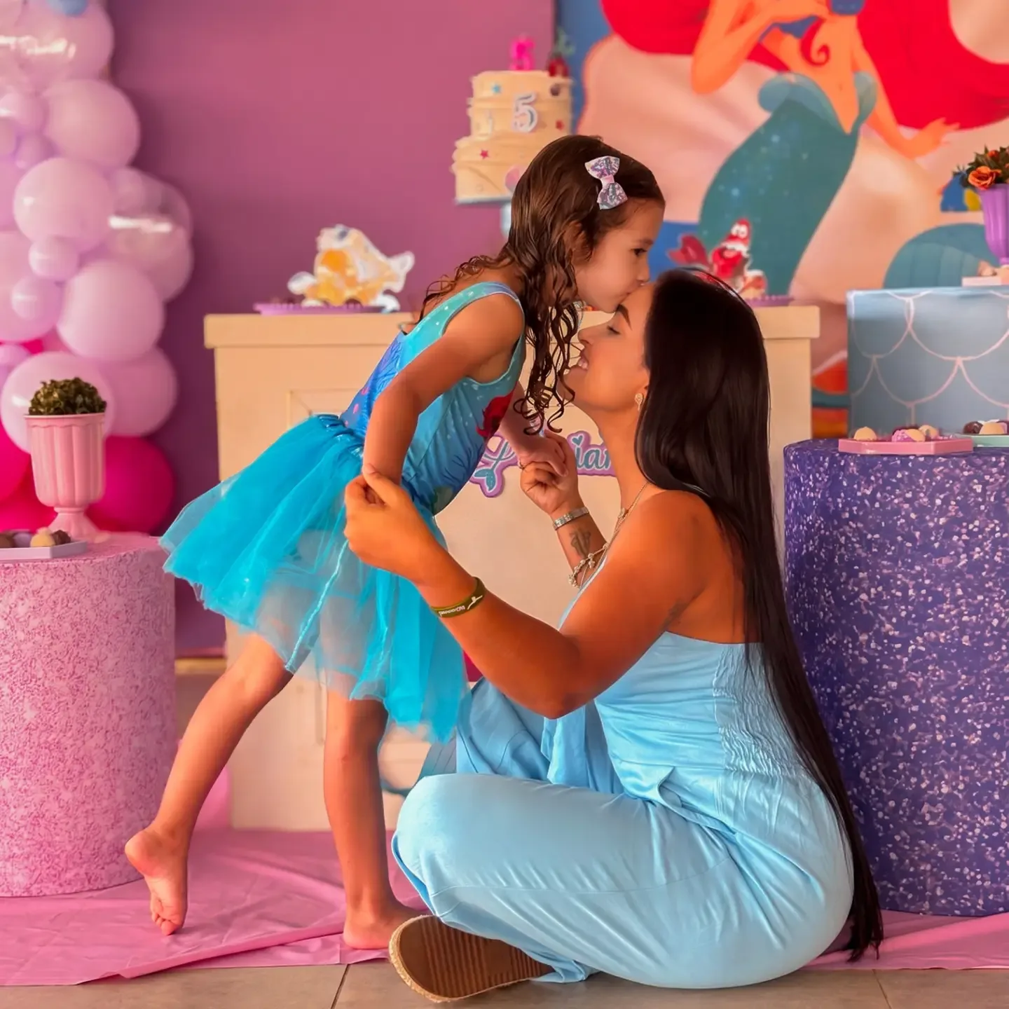 Mulher ajoelhada beija uma menina de vestido azul diante de uma decoração de festa rosa.
