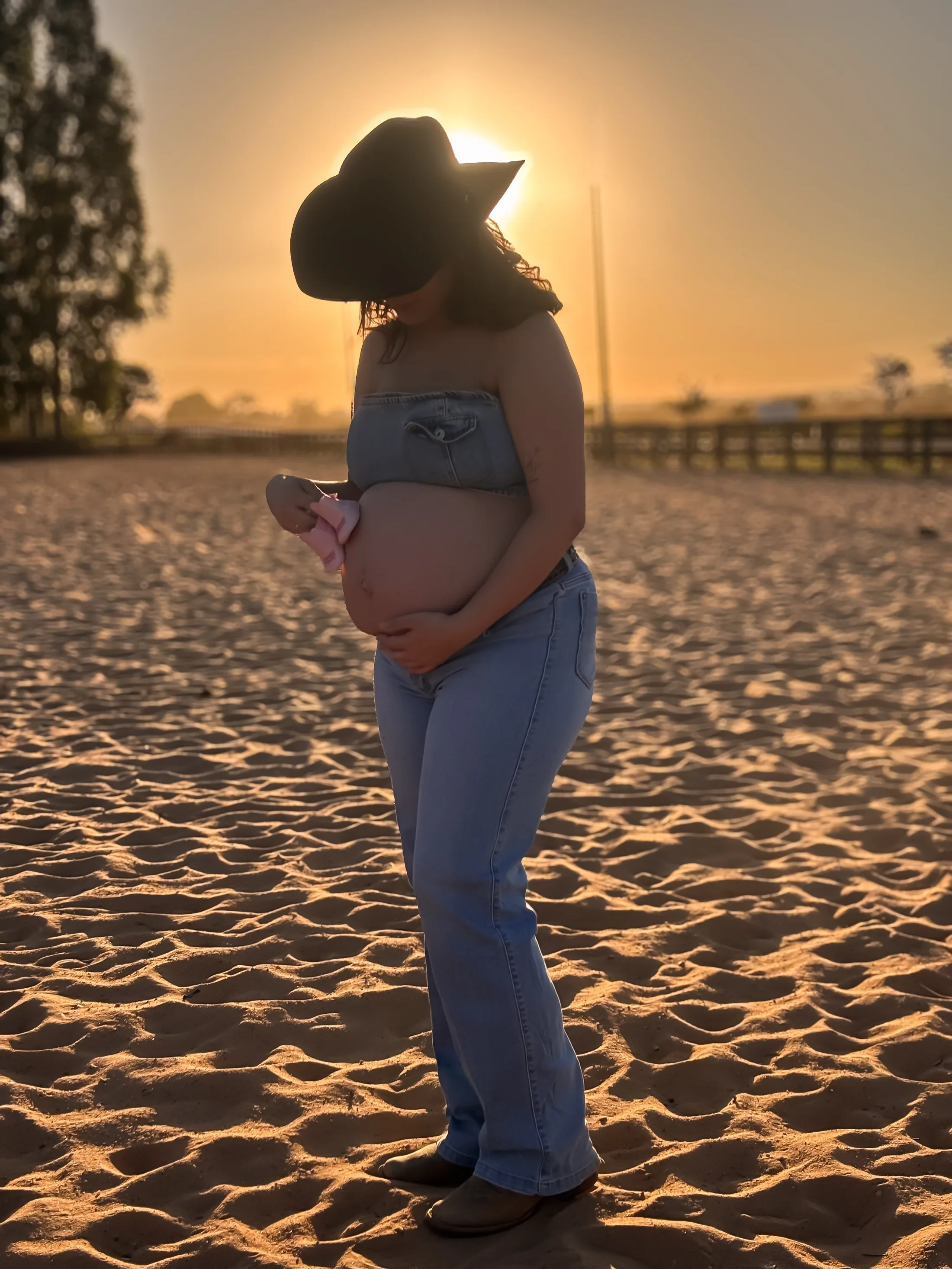 Mulher de chapéu apoia as mãos na barriga em um terreno de areia ao pôr do sol.