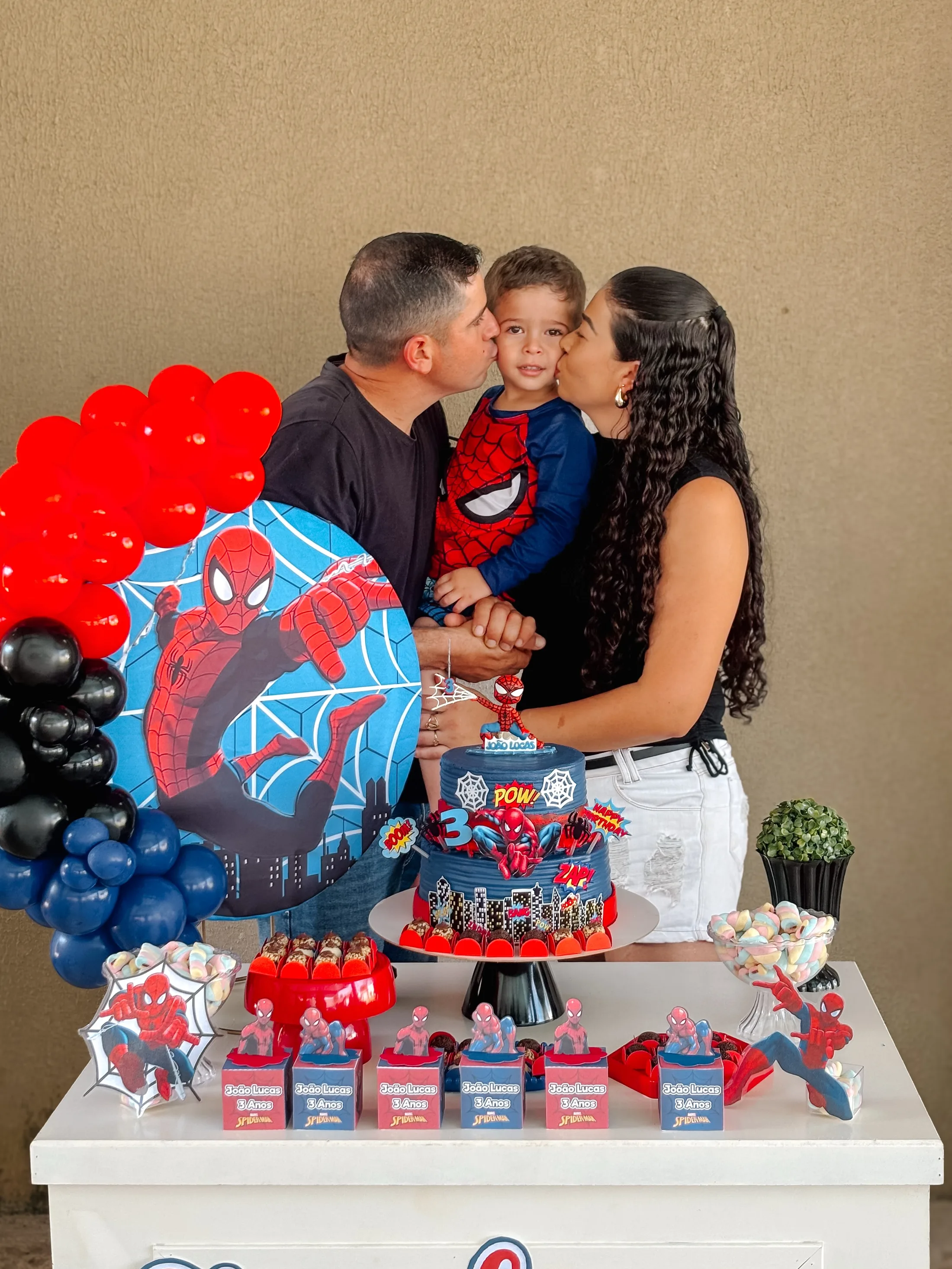 Casal beija uma criança diante de uma mesa decorada em vermelho e azul.
