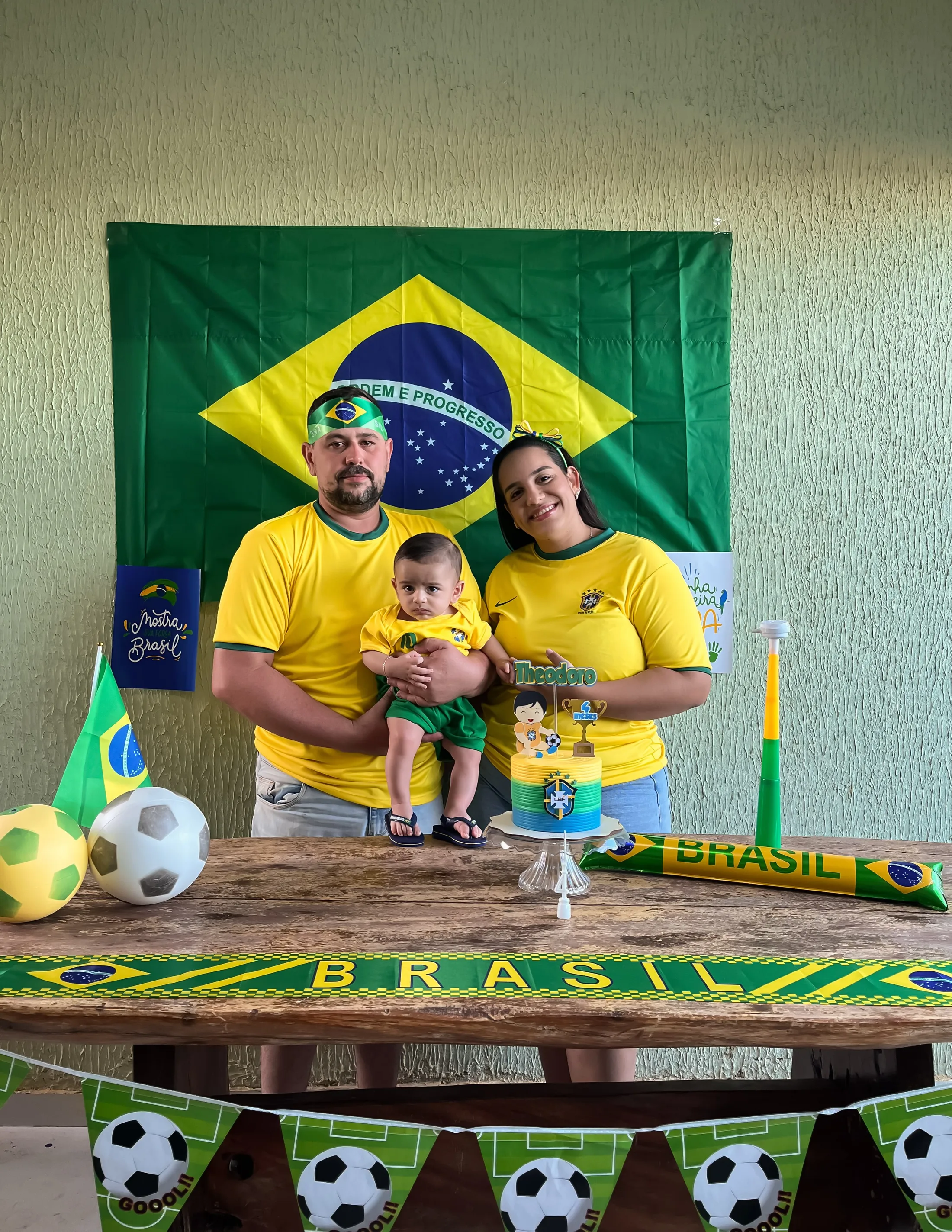 Três pessoas de camisetas amarelas posam atrás de uma mesa decorada com o tema Brasil.