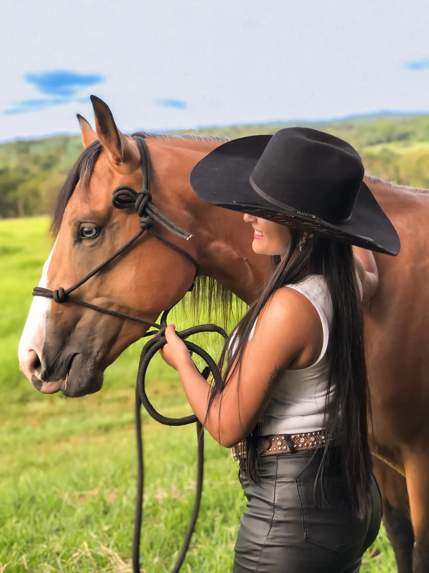 Mulher de chapéu preto junto ao rosto de um cavalo diante de um campo verde.