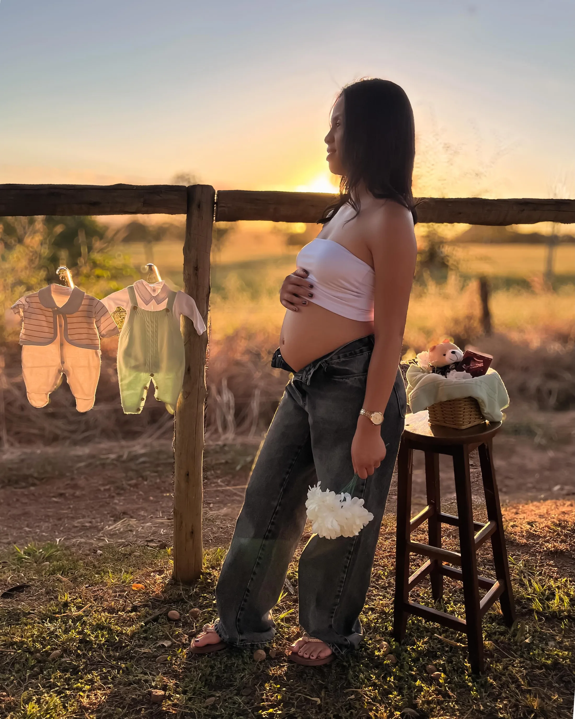 Mulher de top branco apoia as mãos na barriga diante de um varal com pequenas roupas ao entardecer.