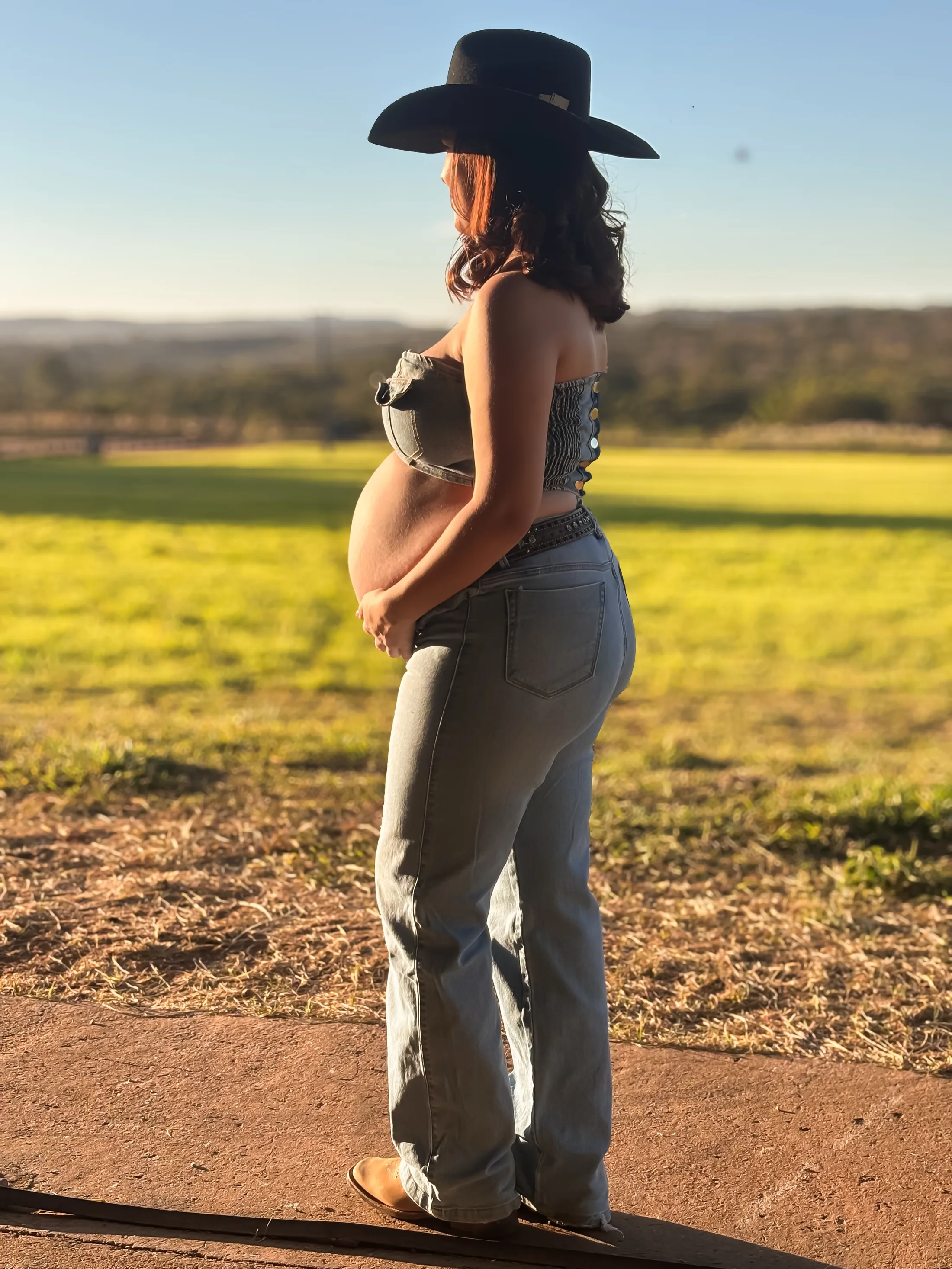 Mulher de chapéu e roupa escura posa de perfil diante de um campo verde ao entardecer.
