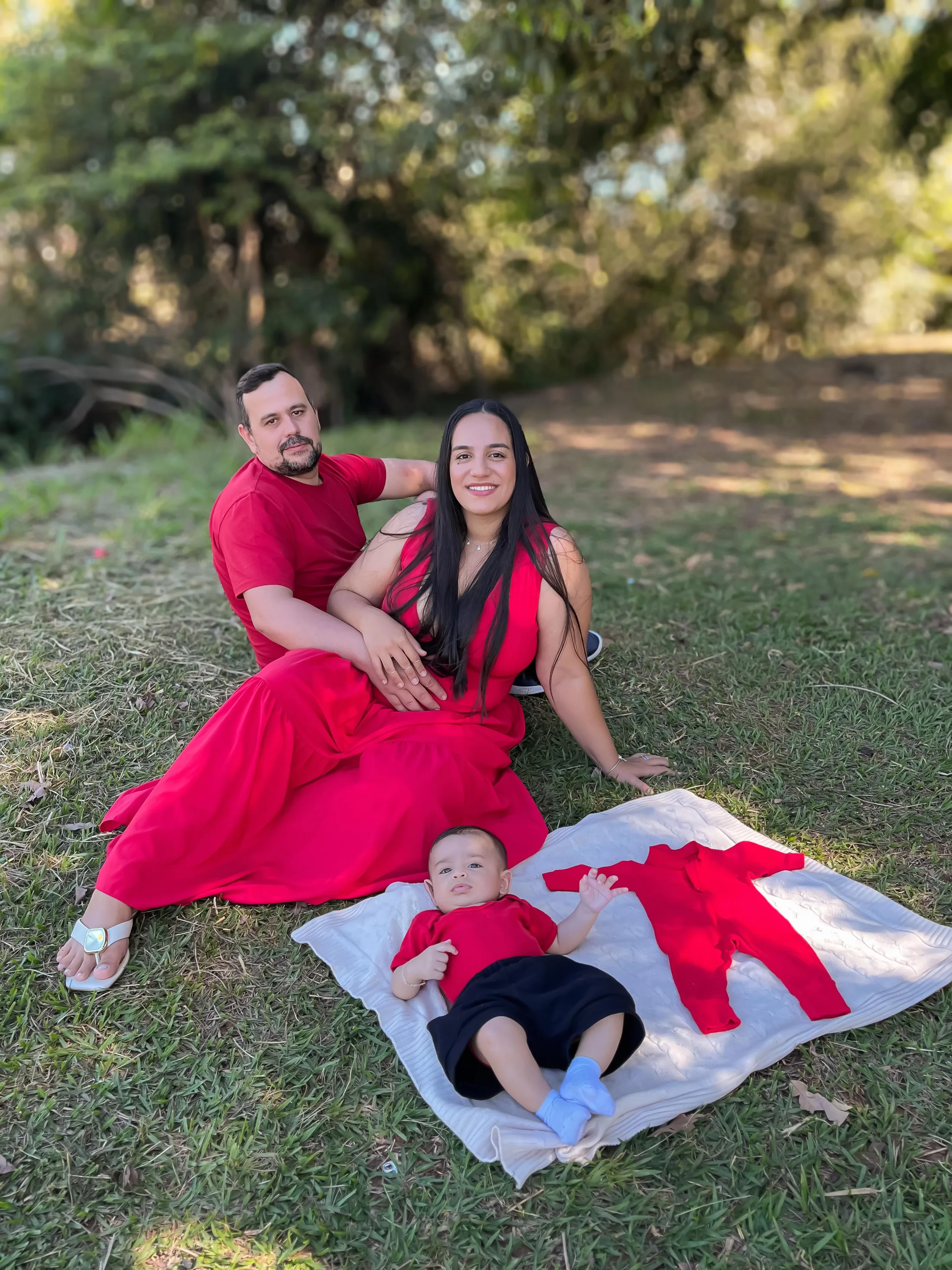 Casal de vermelho sentado na grama, com um bebê deitado sobre uma manta branca à frente.