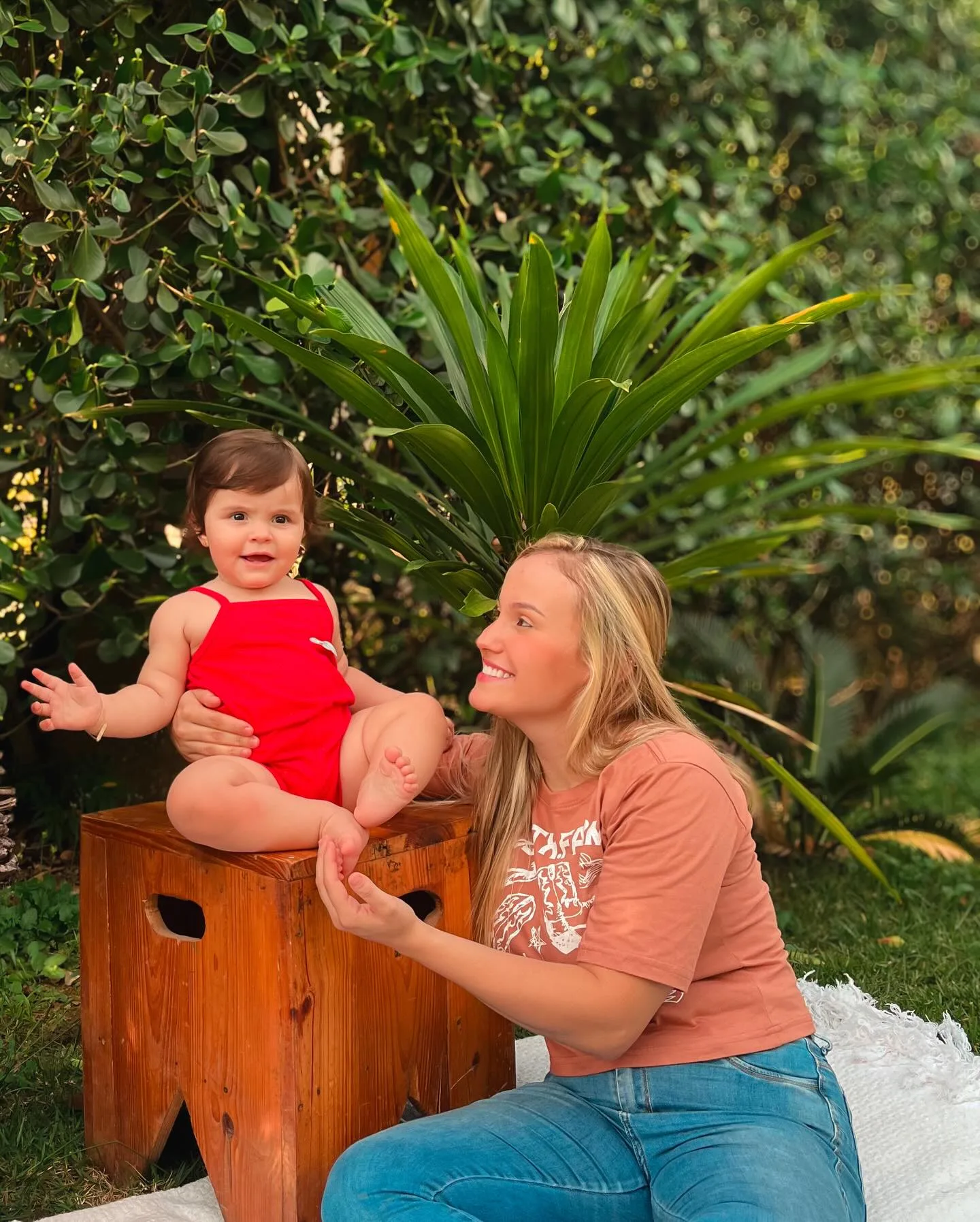 Criança de vermelho sentada sobre um banco junto a uma mulher em um jardim.
