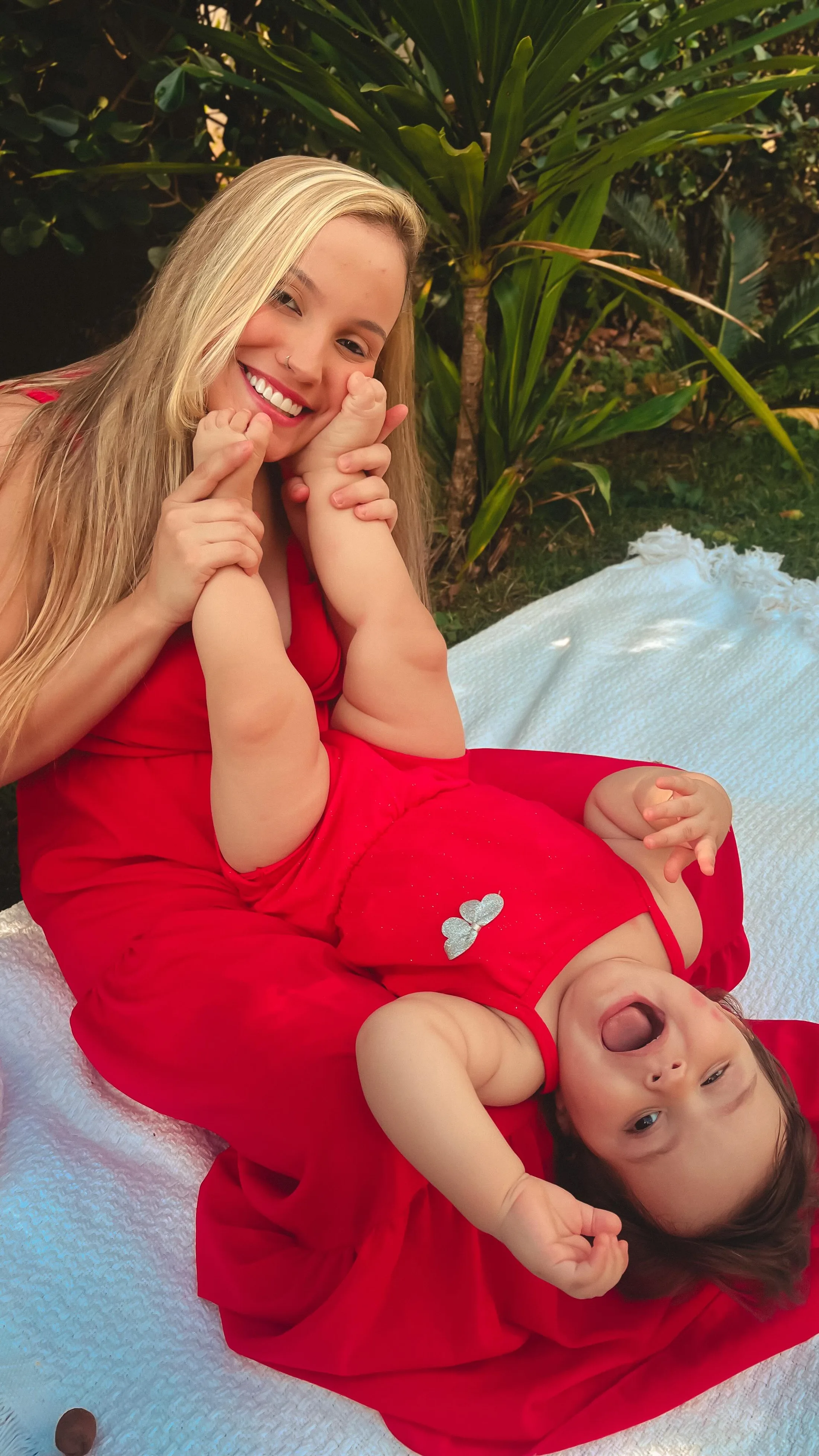 Mulher de vestido vermelho e criança de vermelho brincam deitadas sobre um tecido branco ao ar livre.