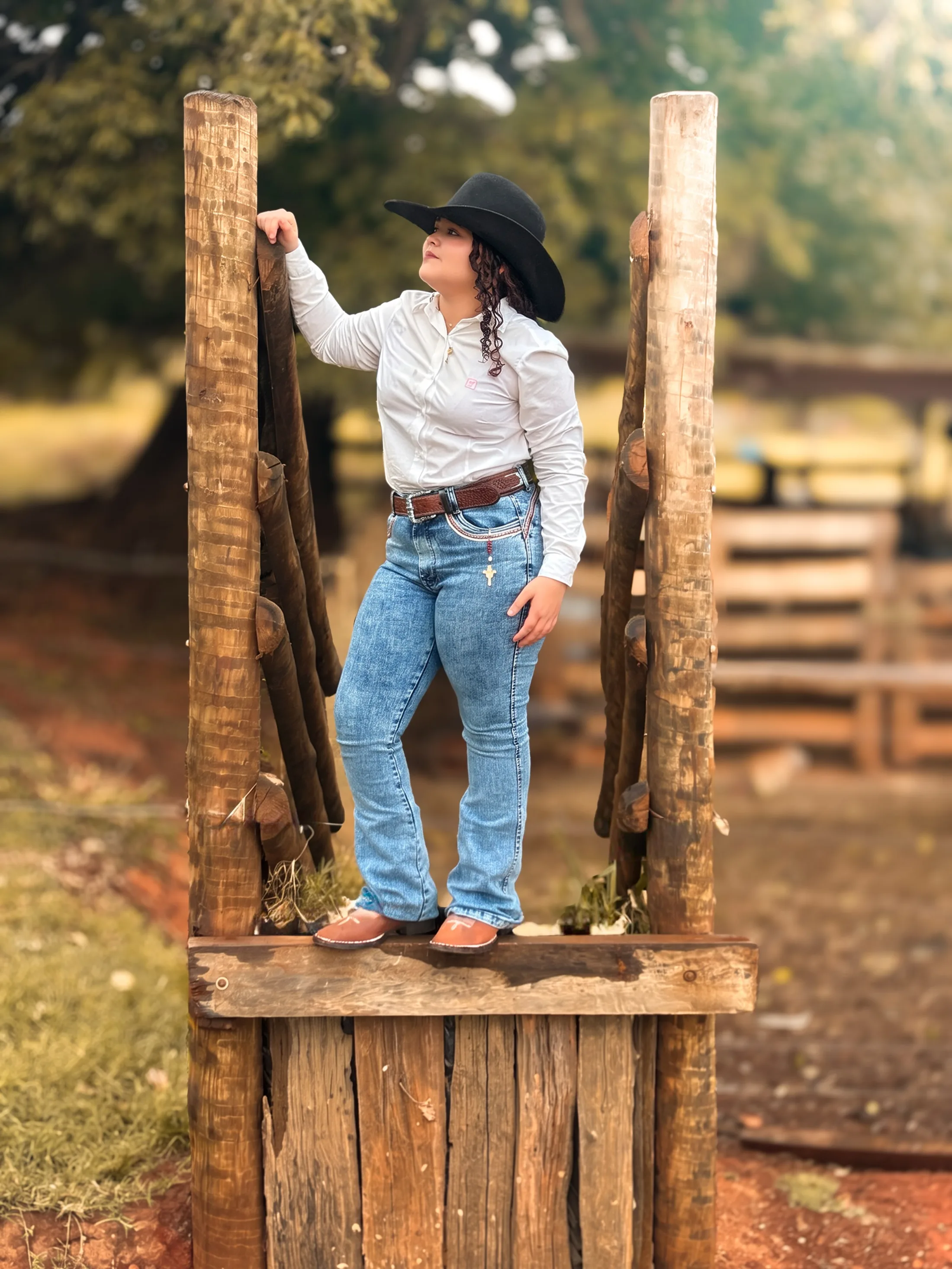 Mulher de chapéu, camisa branca e calça jeans posa sobre uma estrutura de madeira ao ar livre.