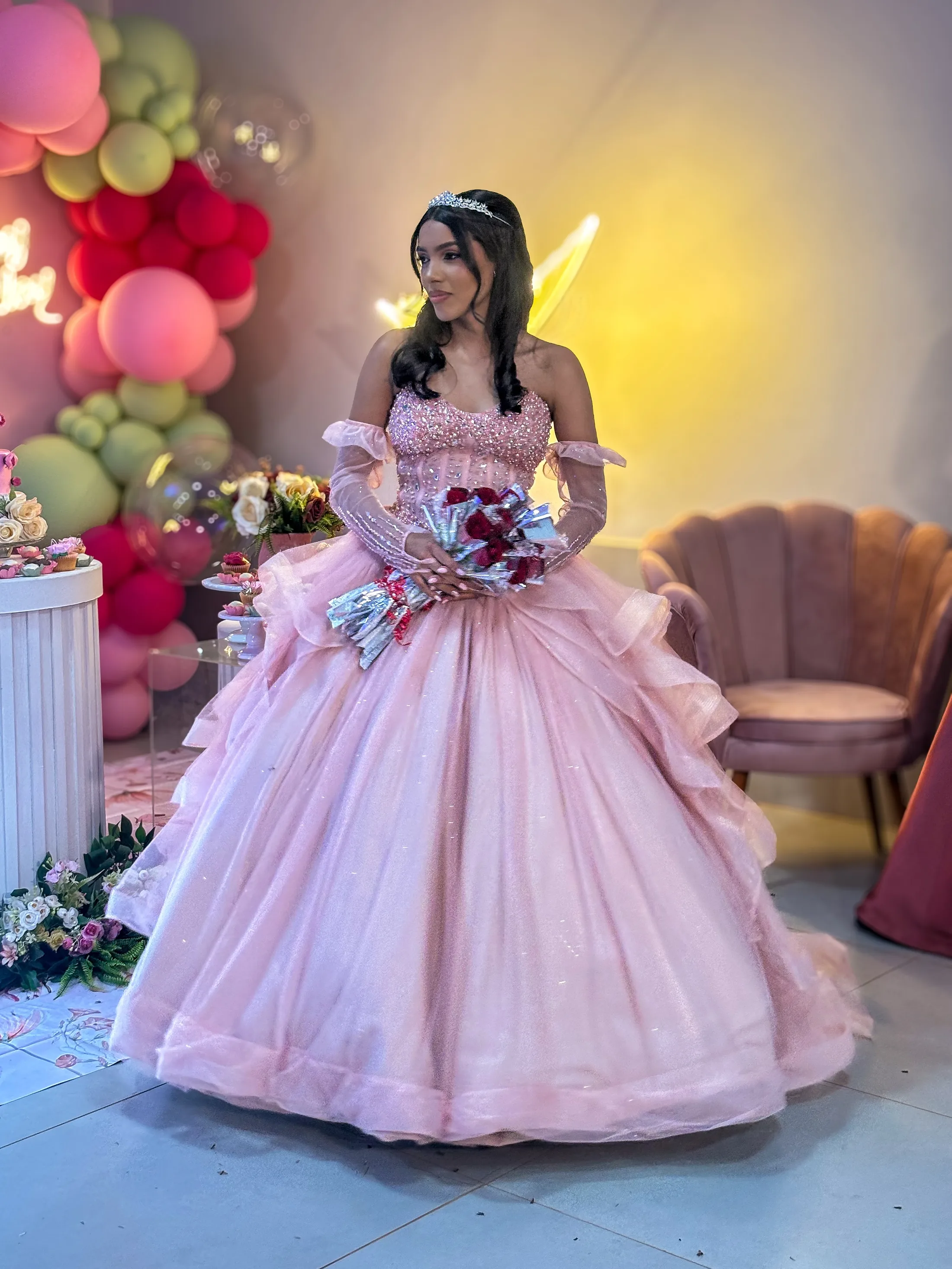 Mulher de vestido rosa amplo segura um buquê diante de uma decoração de festa iluminada.