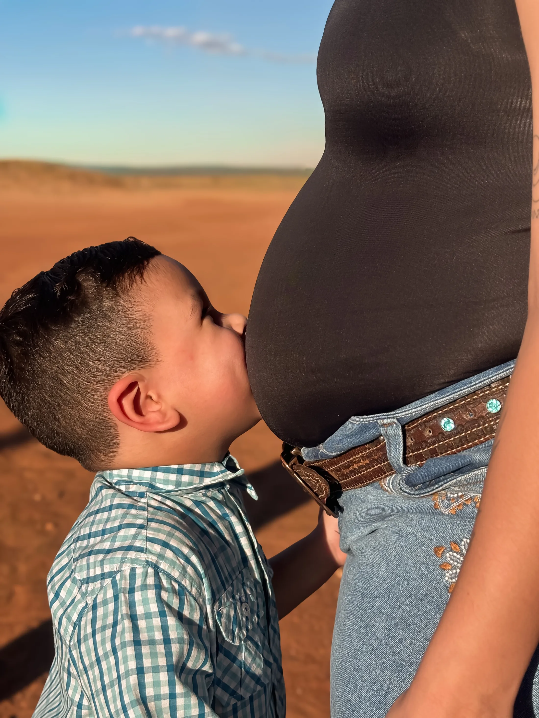 Menino de camisa xadrez beija uma barriga de gestante em um cenário ao ar livre.