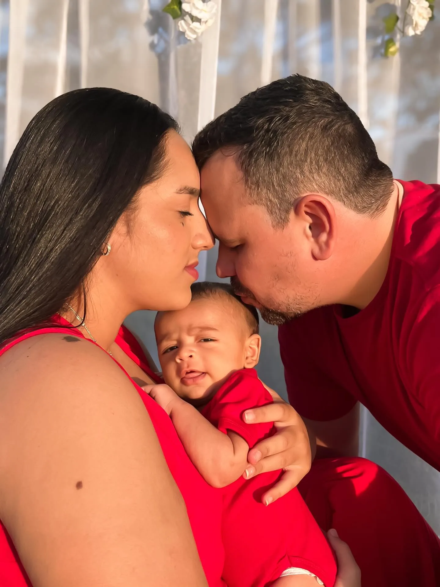 Casal de vermelho aproxima o rosto enquanto segura um bebê de vermelho em um jardim.