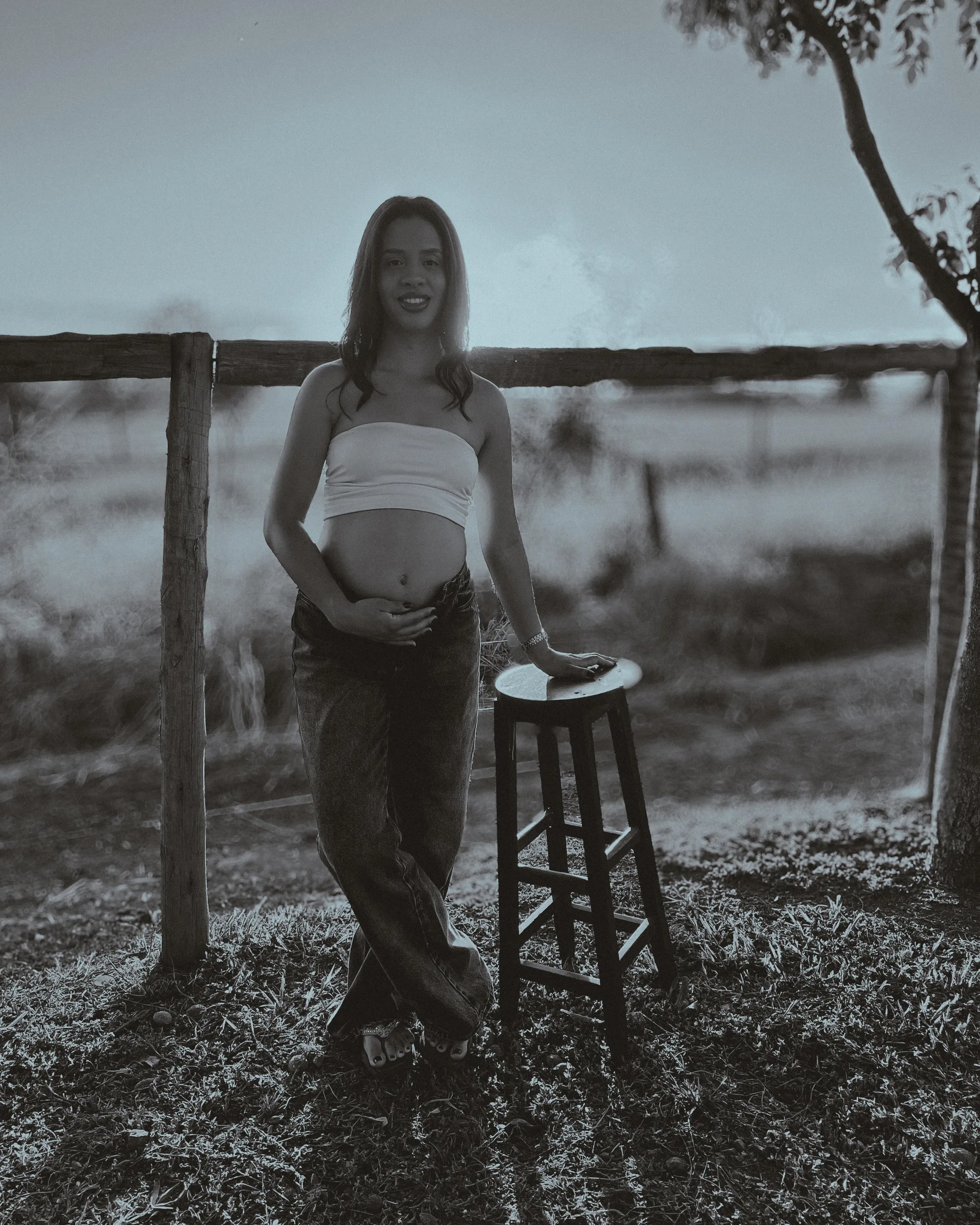 Mulher posa junto a uma cerca e a um banco em fotografia de gestação em preto e branco.