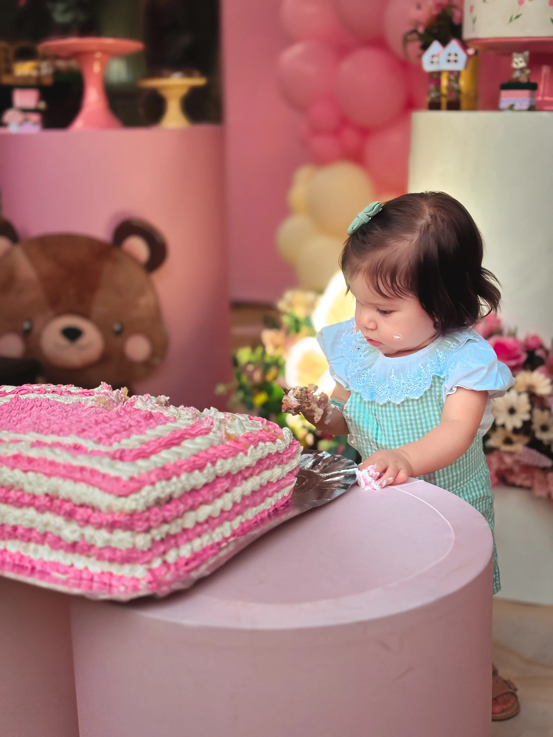 Menina de vestido azul claro olha para um bolo rosa sobre uma mesa de festa.