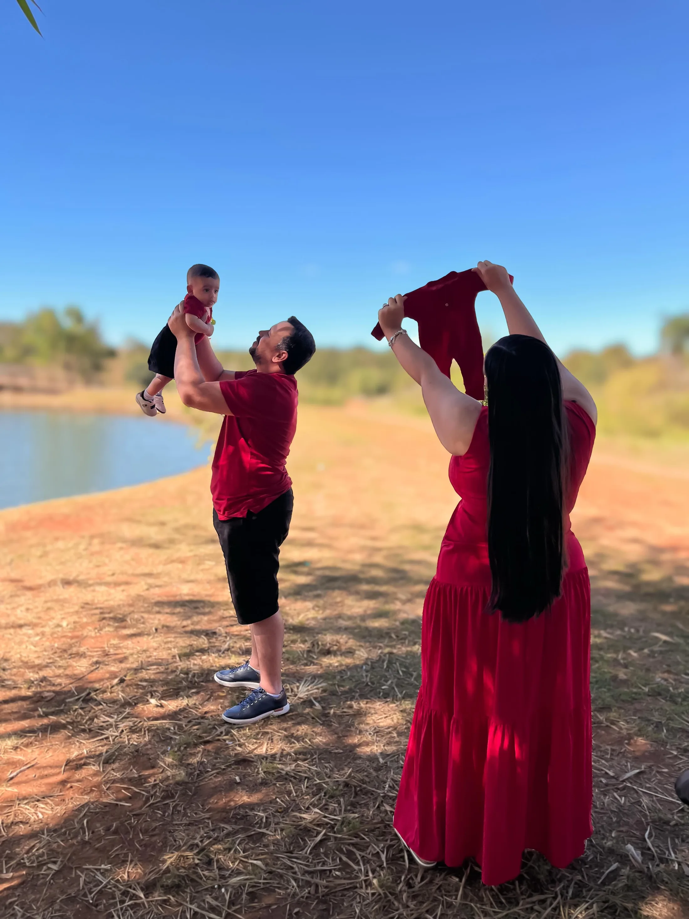 Casal de vermelho ergue uma criança junto a um lago sob o céu azul.