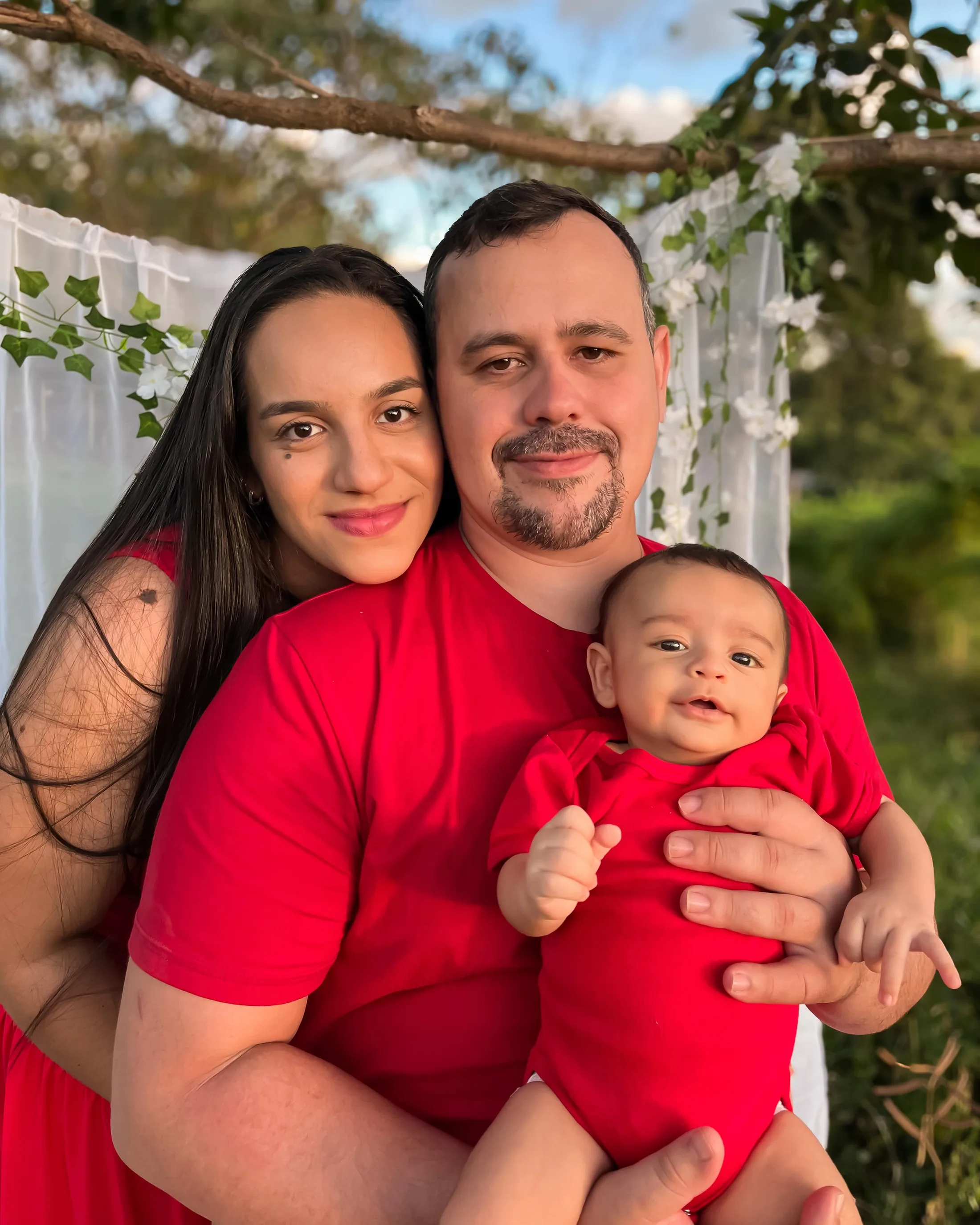 Mulher e homem de vermelho seguram um bebê diante de um cenário branco ao ar livre.