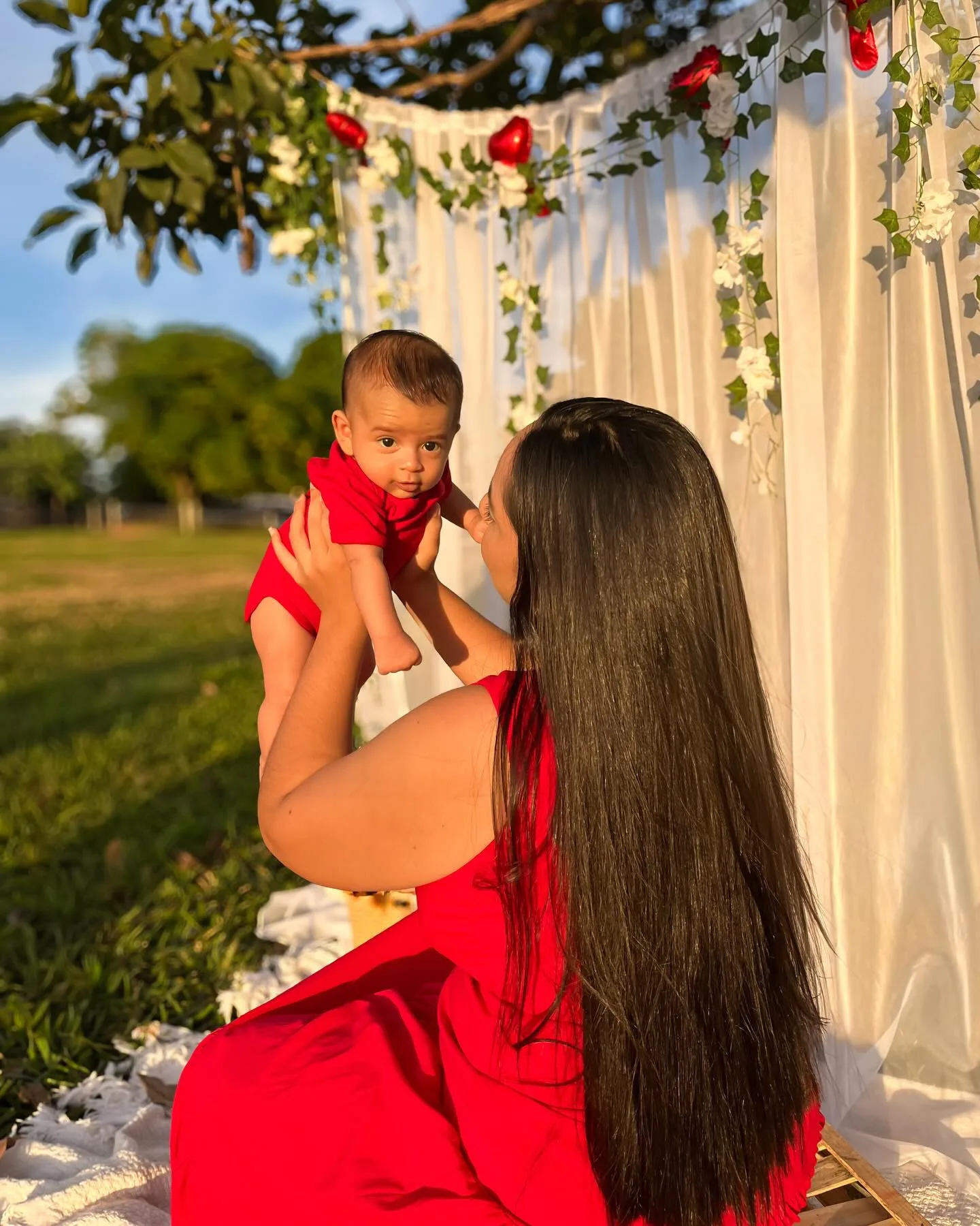 Mulher de vermelho ergue uma criança diante de um cenário branco com flores.