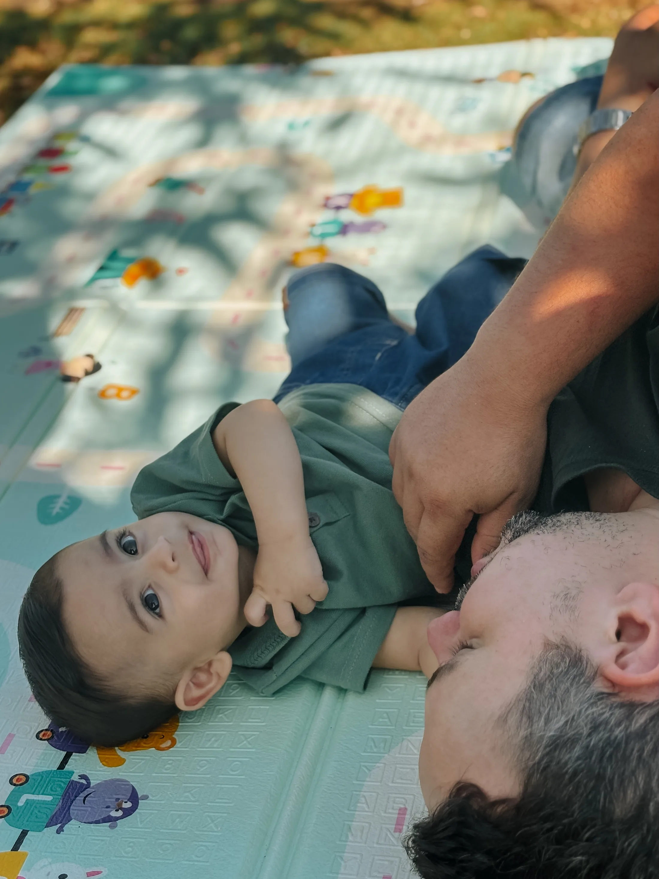 Bebê de roupa verde deitado sobre uma manta junto a um homem ao ar livre.