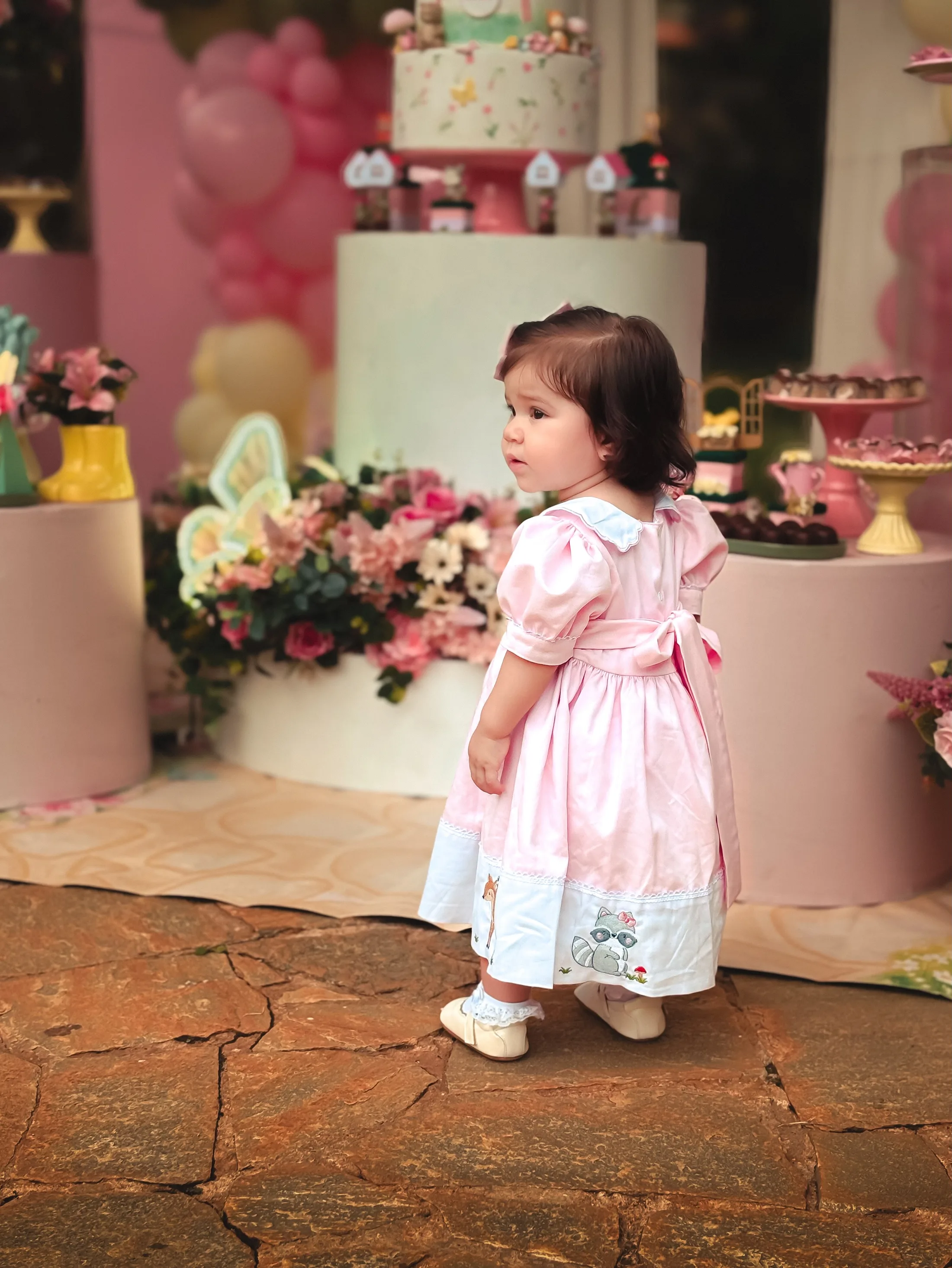 Menina de vestido rosa claro em pé diante de uma decoração com flores e balões.