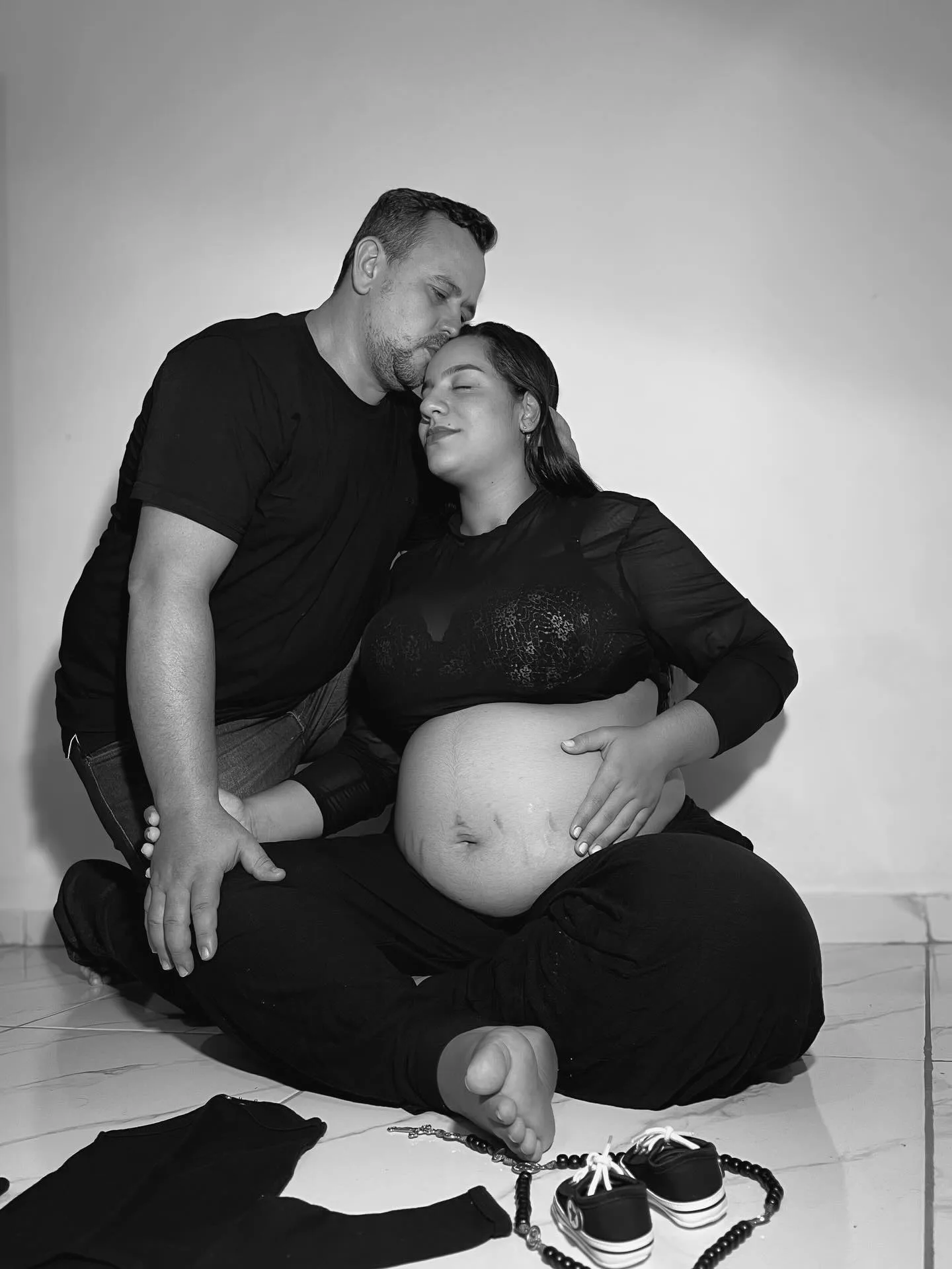 Casal sentado em fundo claro durante ensaio de gestação em preto e branco.