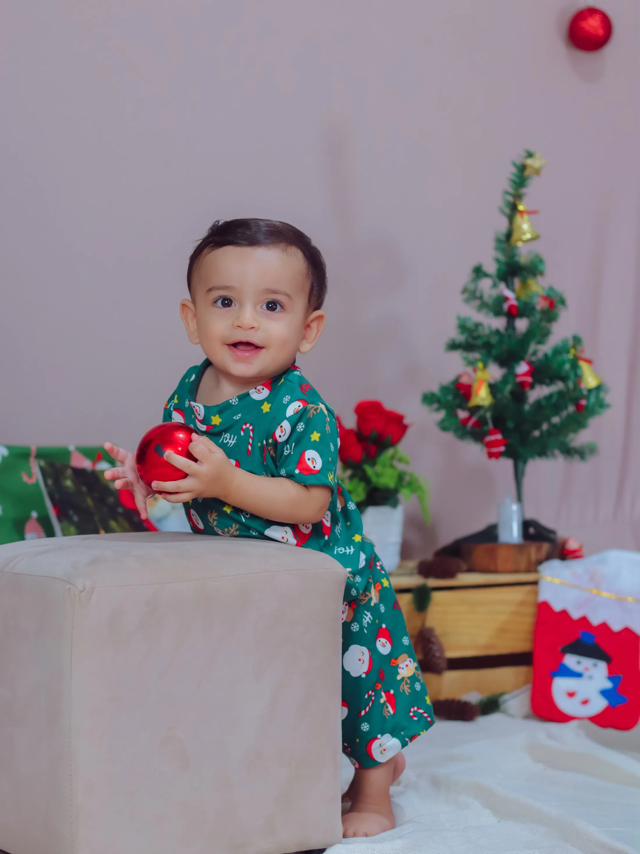 Bebê de roupa estampada verde junto a uma pequena árvore de Natal.