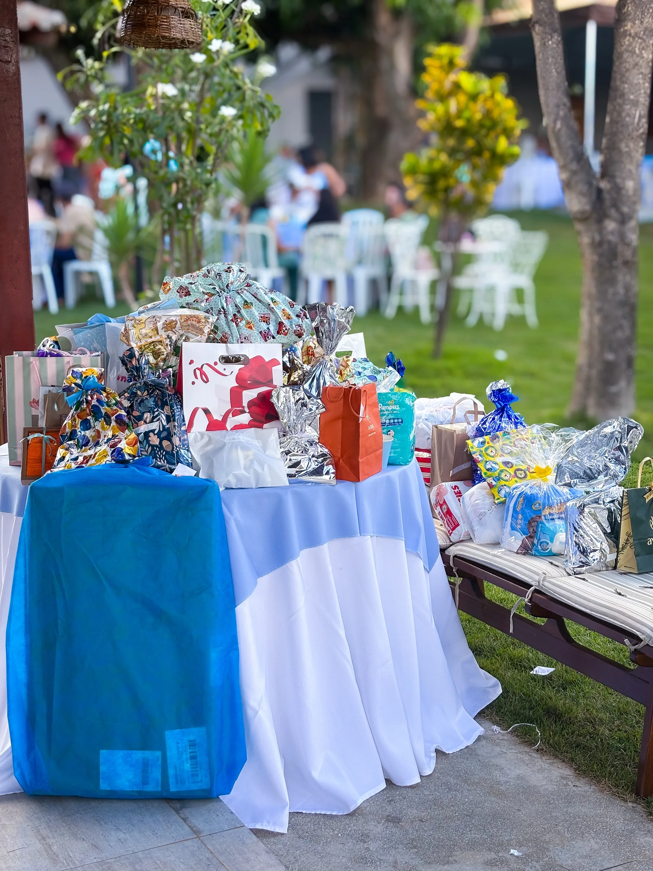 Mesas cobertas com tecidos azul e branco exibem doces e lembranças em um jardim.