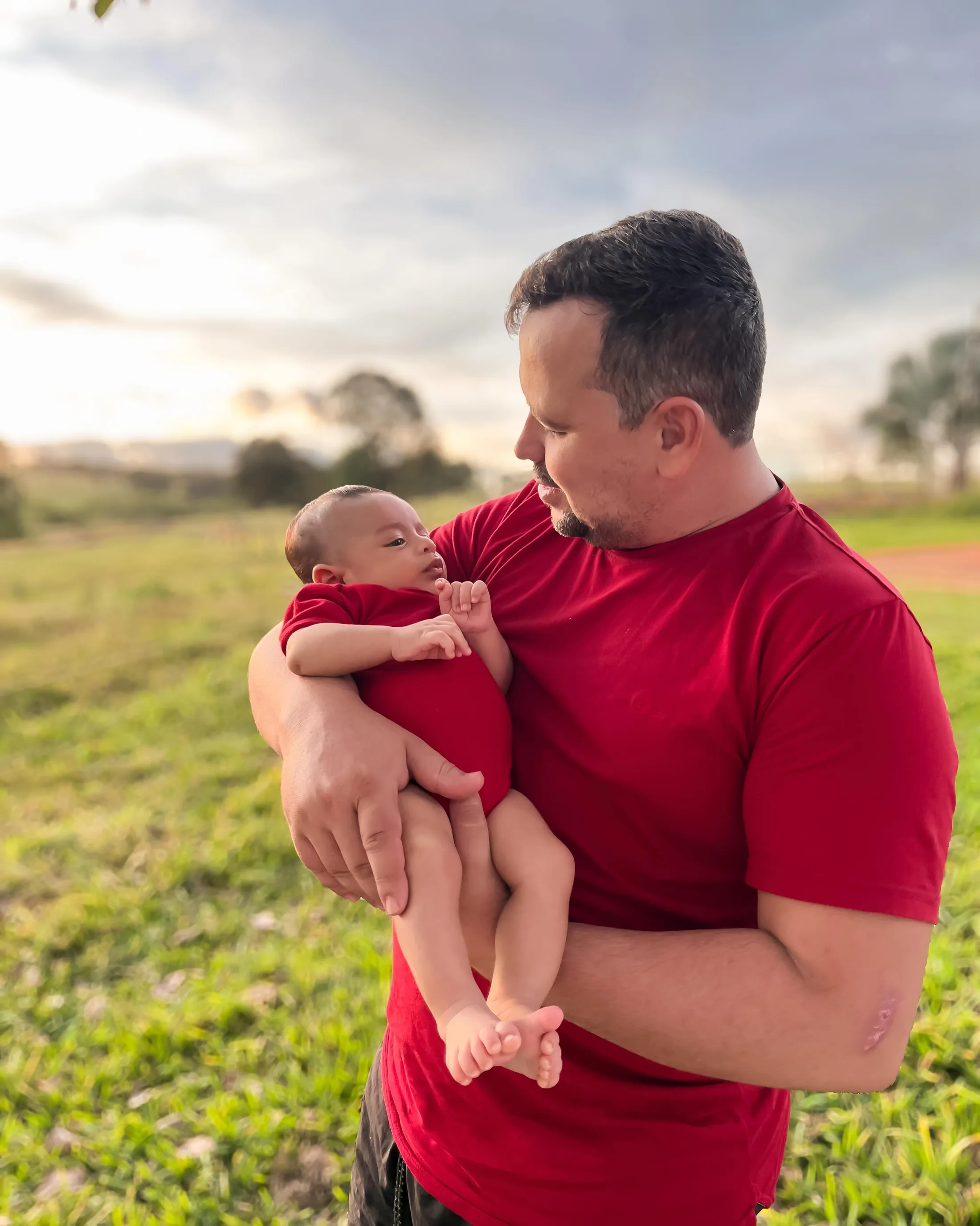 Homem de camiseta vermelha segura um bebê de vermelho em um campo ao entardecer.