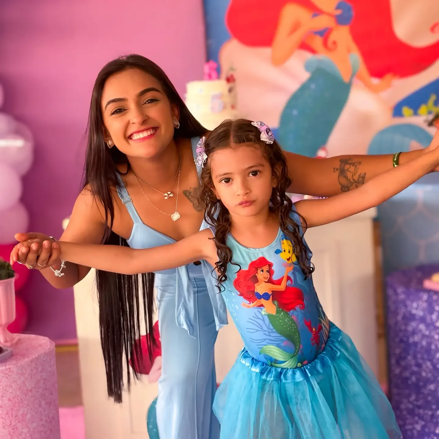 Mulher e menina de vestido azul sorriem diante de uma decoração de festa rosa.