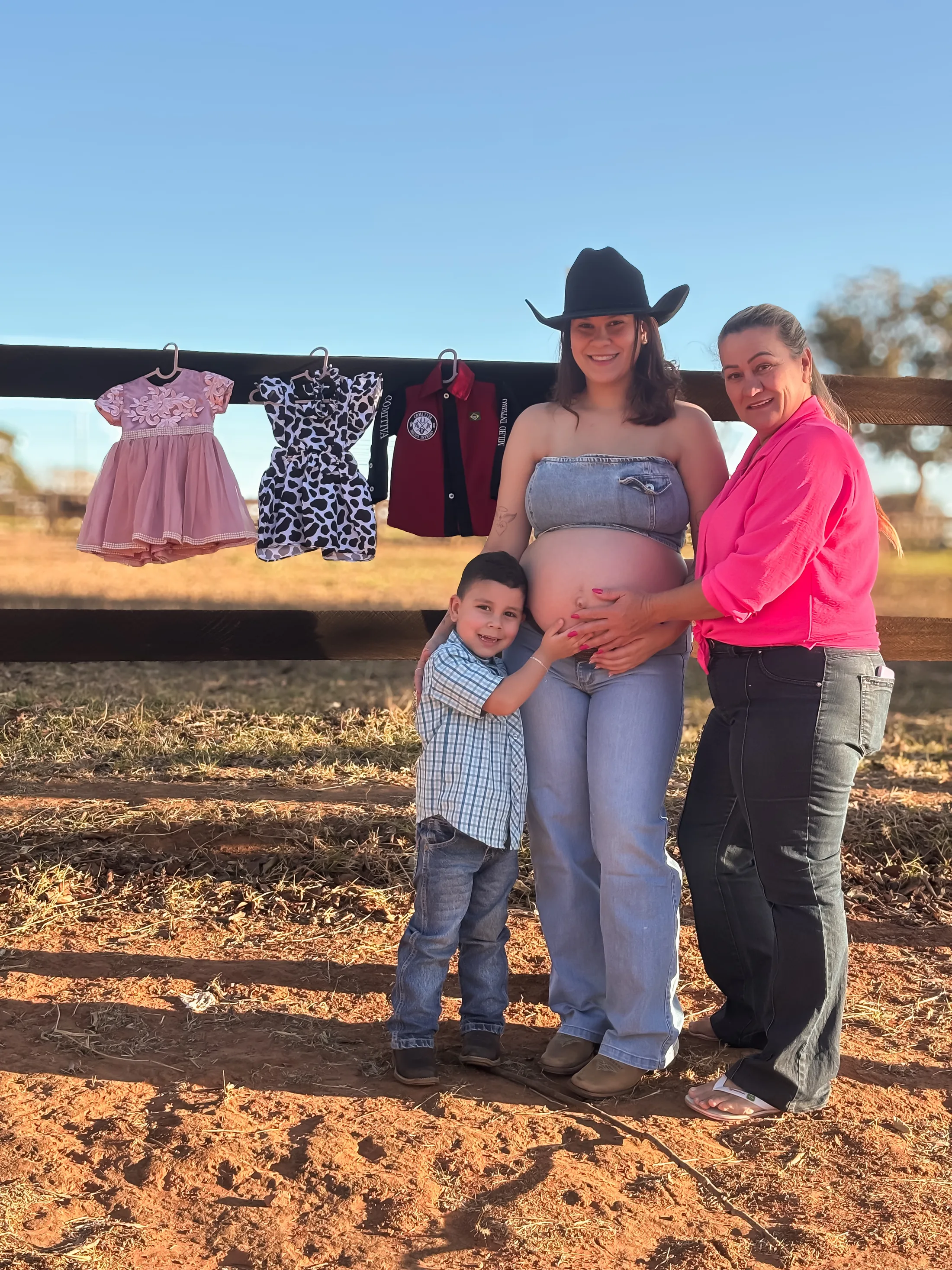 Mulheres e menino posam junto a um varal de pequenas roupas em um cenário rural.