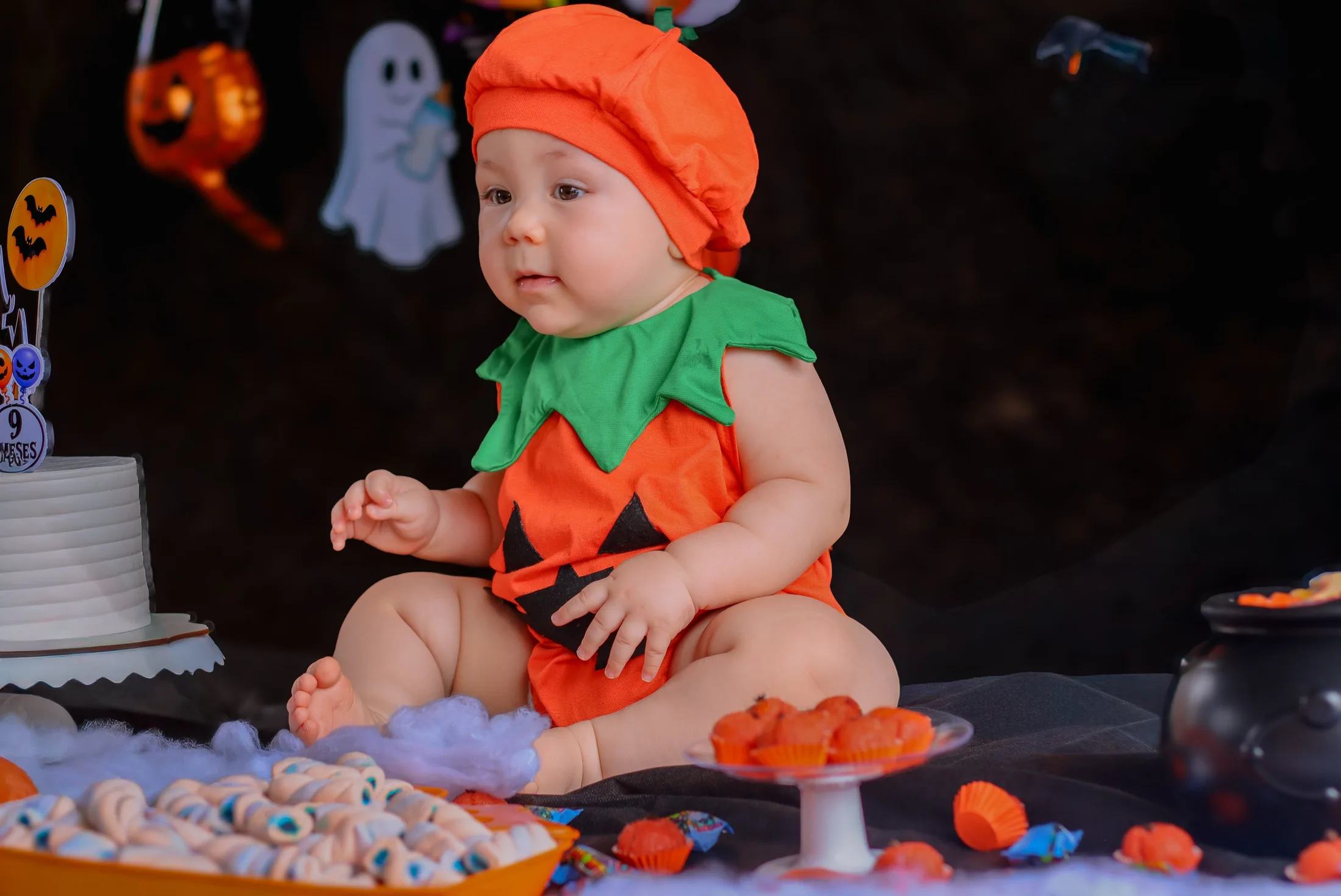 Bebê com roupa de abóbora sentado diante de um cenário escuro com pequenos fantasmas.