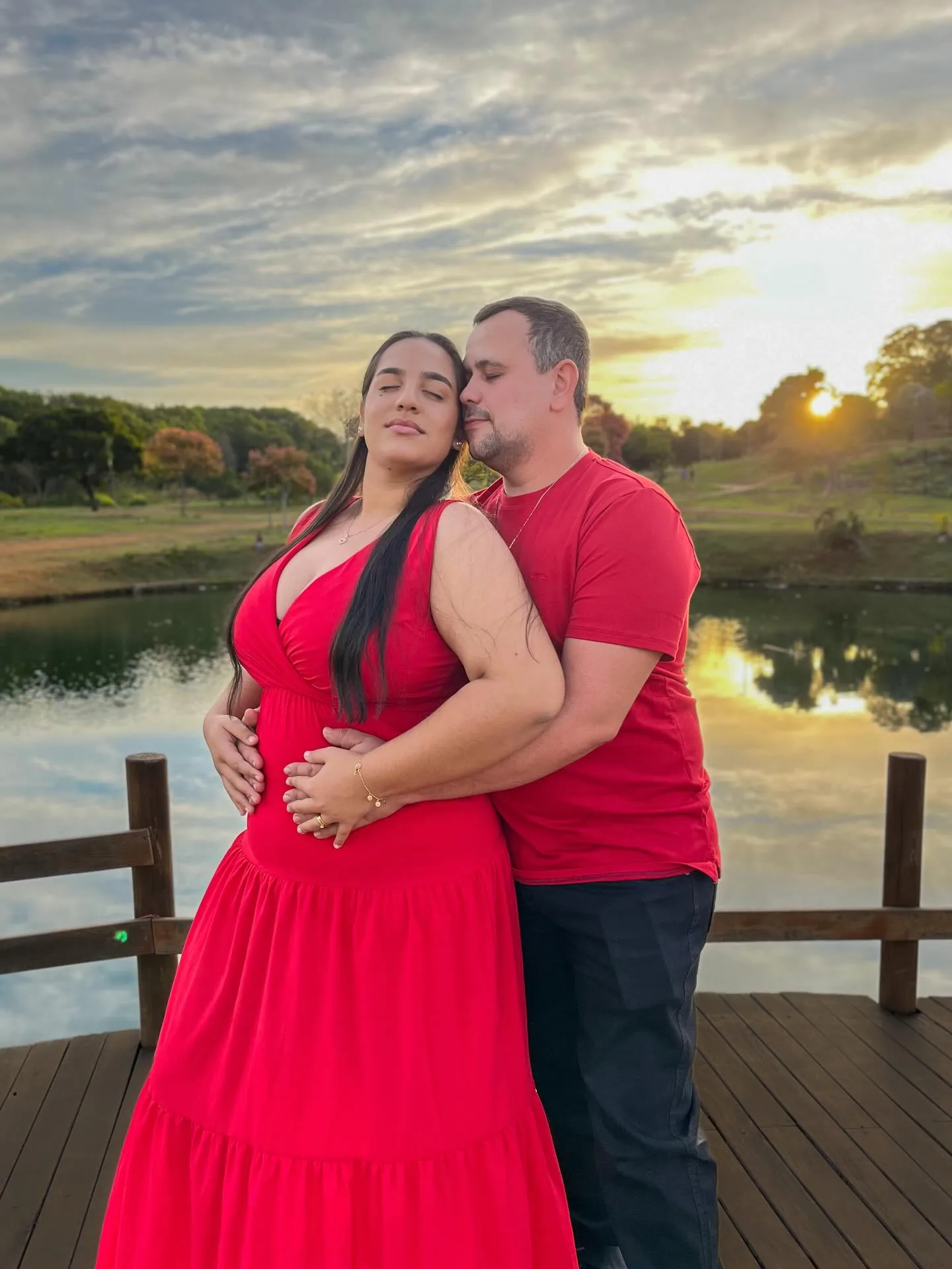 Casal de vermelho se abraça sobre uma ponte de madeira, diante de um lago ao entardecer.