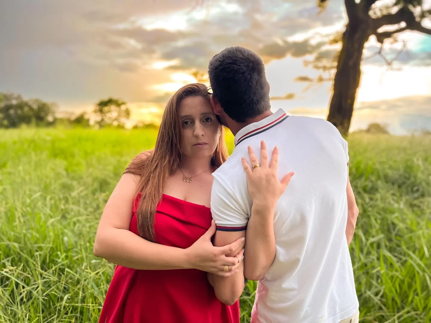 Casal abraçado em um campo verde, com vestido vermelho e céu iluminado ao fundo.