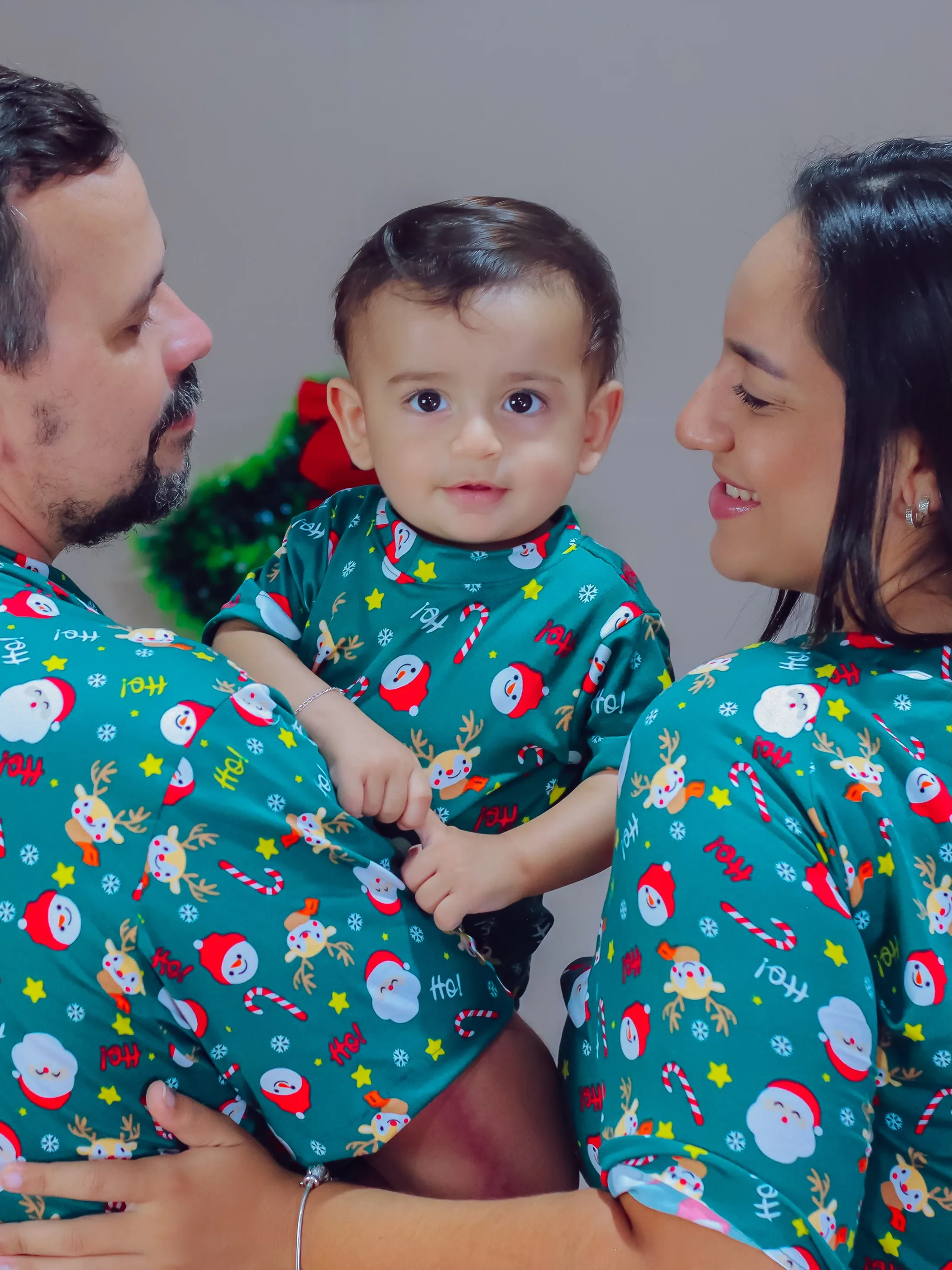 Casal e bebê de pijamas verdes estampados posam juntos diante de uma cortina clara.