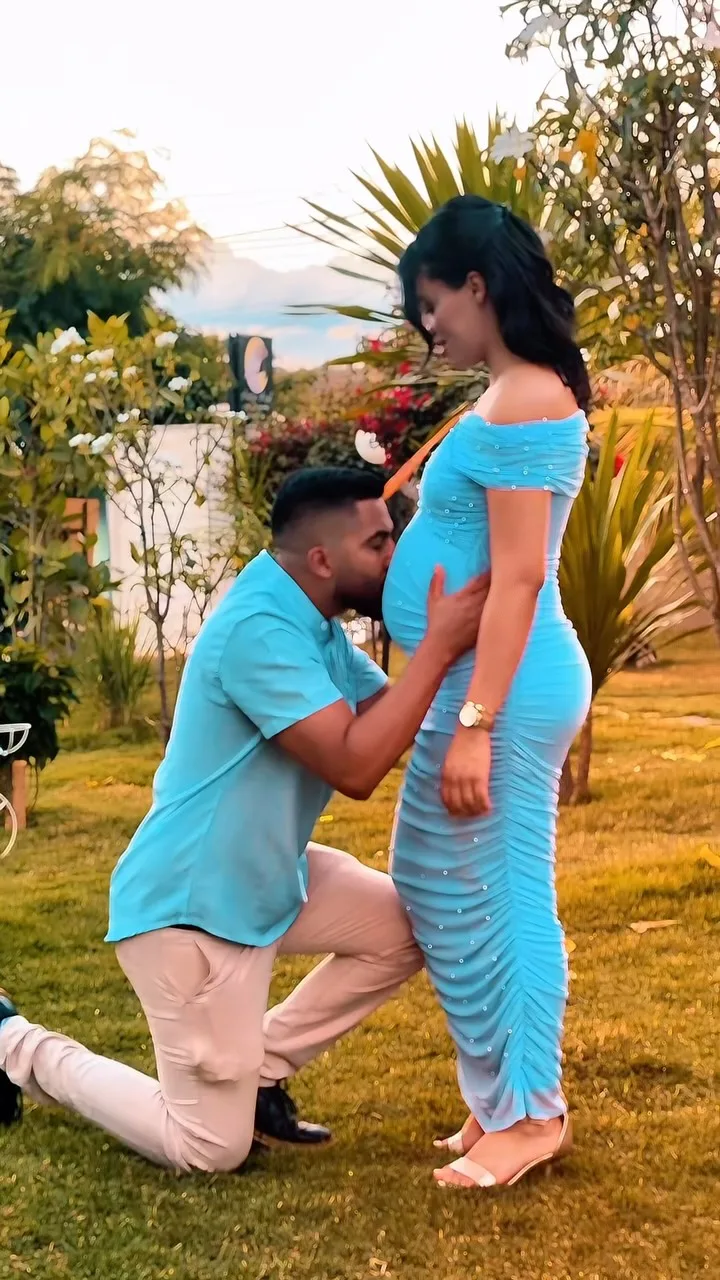 Homem ajoelhado aproxima o rosto da barriga de uma mulher de vestido azul em um jardim.