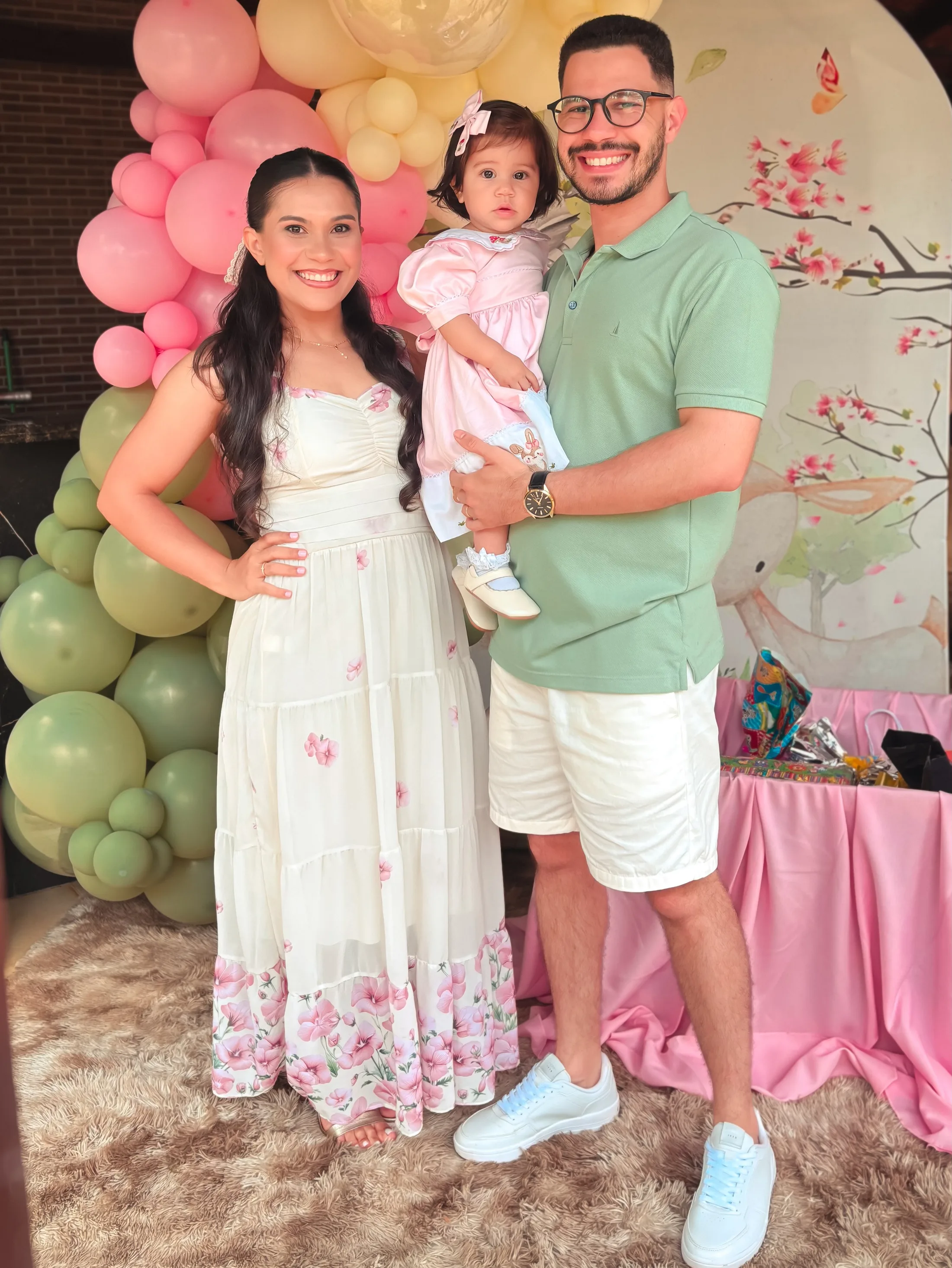 Casal segura uma menina de vestido rosa diante de uma decoração de balões rosas e verdes.