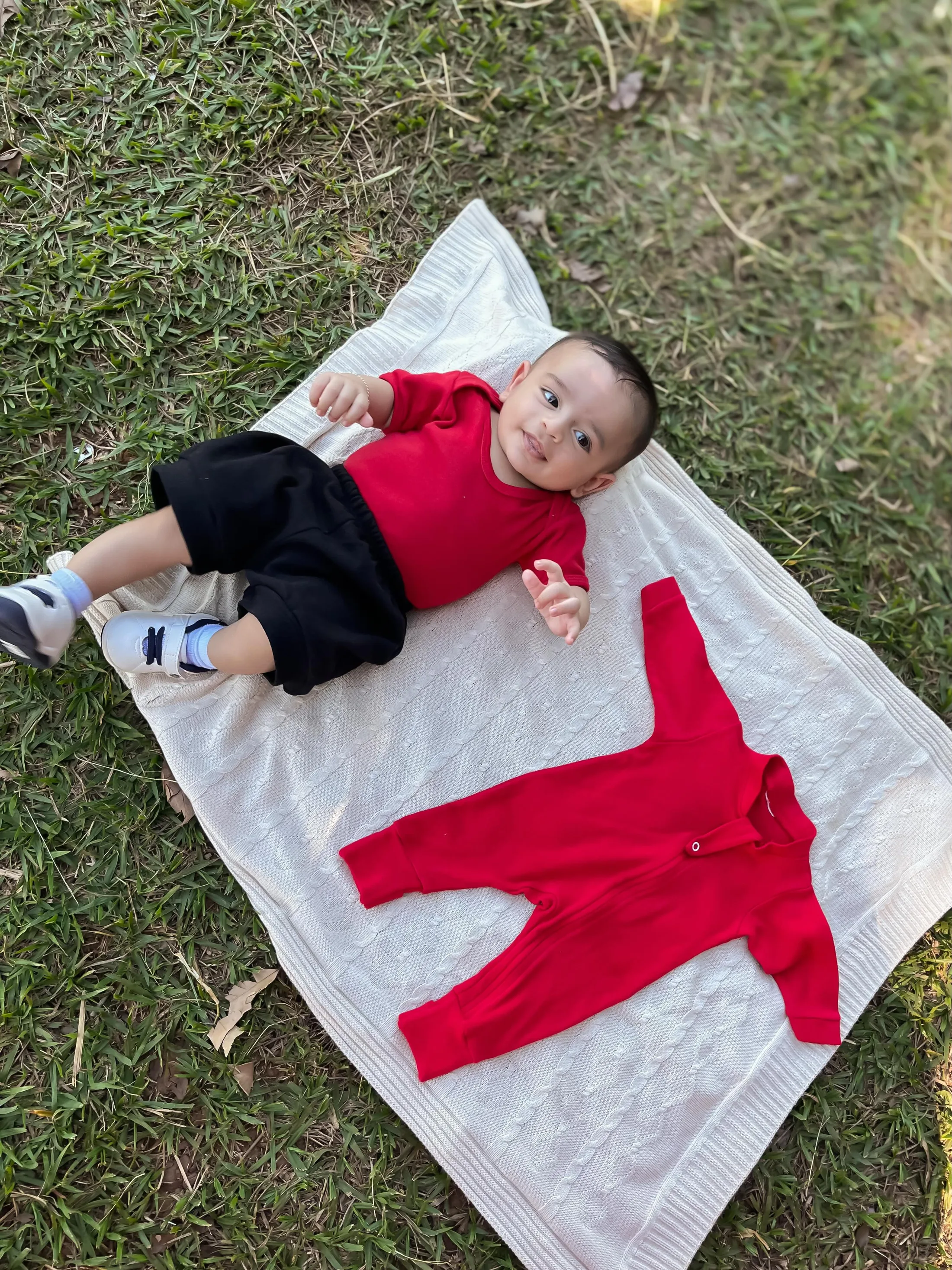 Bebê de camisa vermelha deitado sobre uma manta branca na grama.
