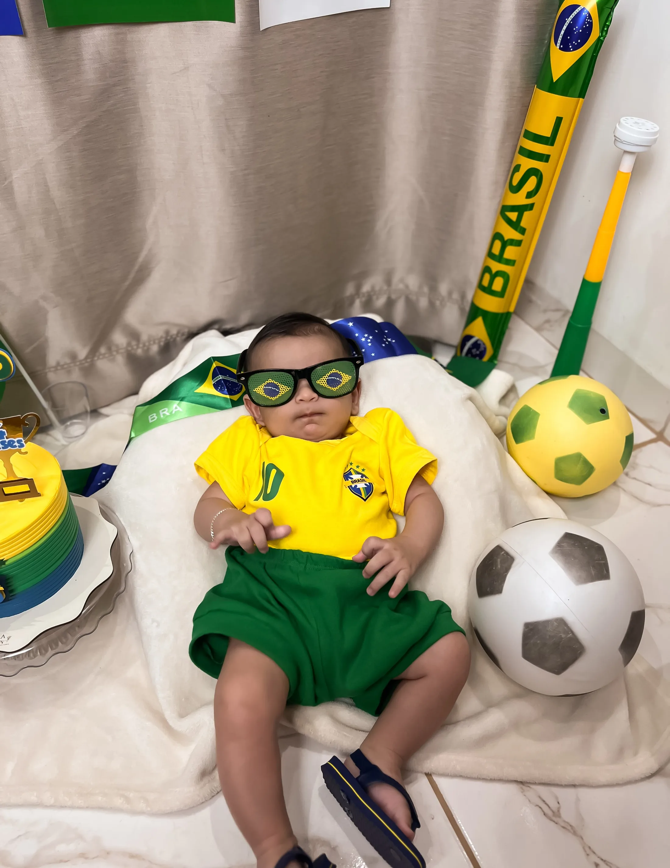 Bebê com óculos escuros e roupa verde e amarela deitado junto a bolas e bandeira do Brasil.