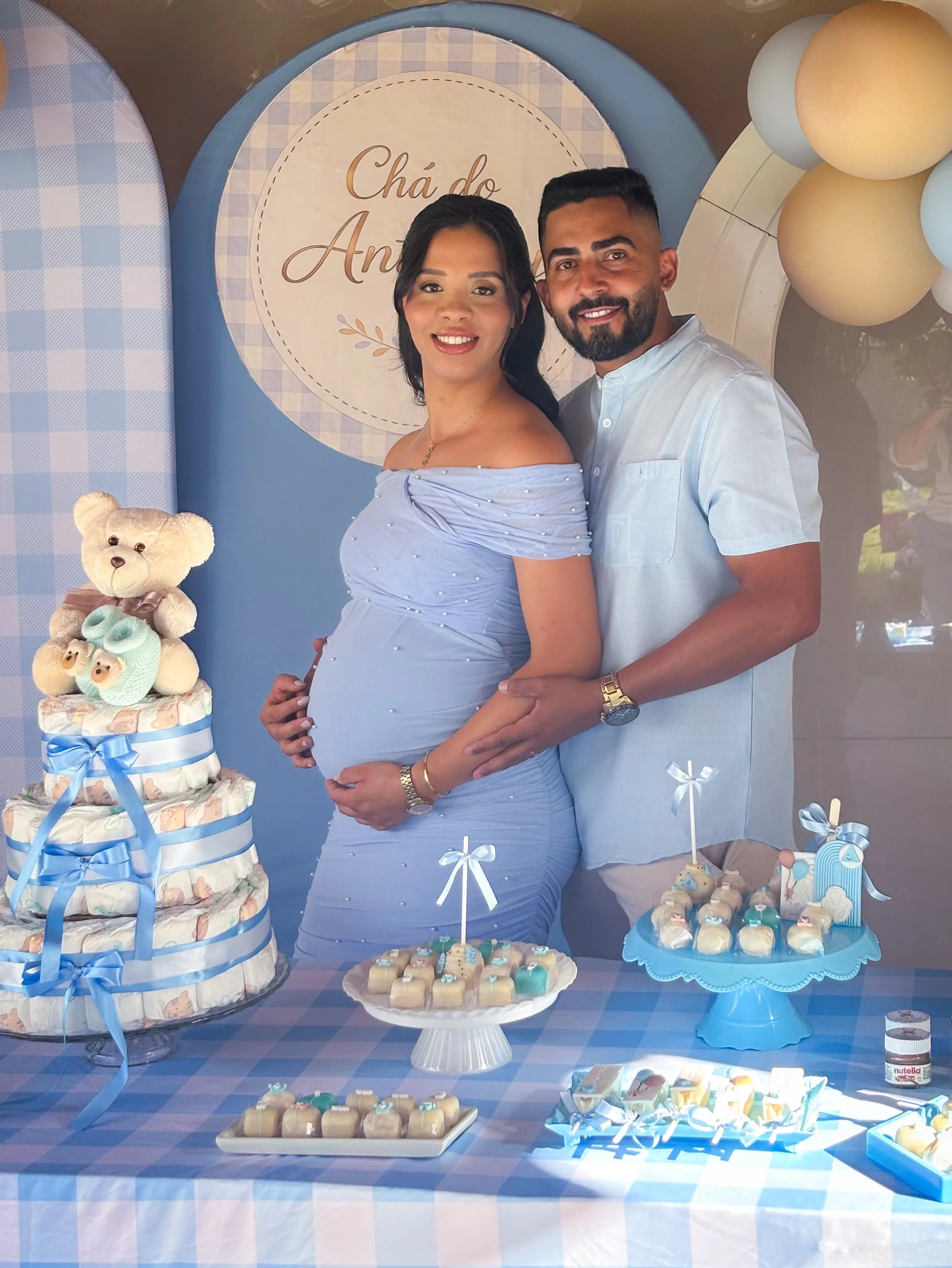 Casal posa atrás de uma mesa azul e branca decorada com um bolo e ursinhos.