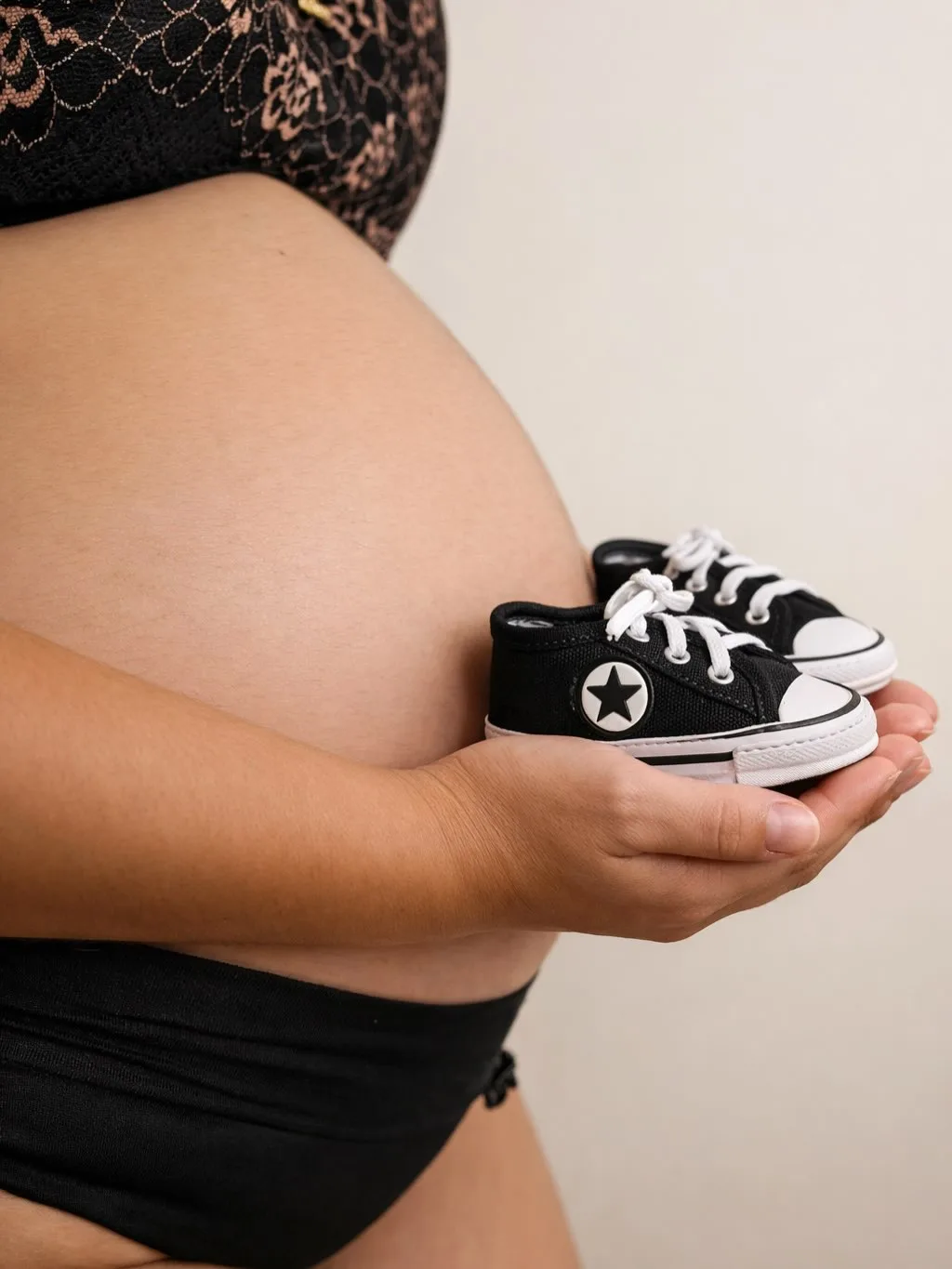 Detalhe de uma barriga de gestante e das mãos que seguram pequenos tênis pretos.