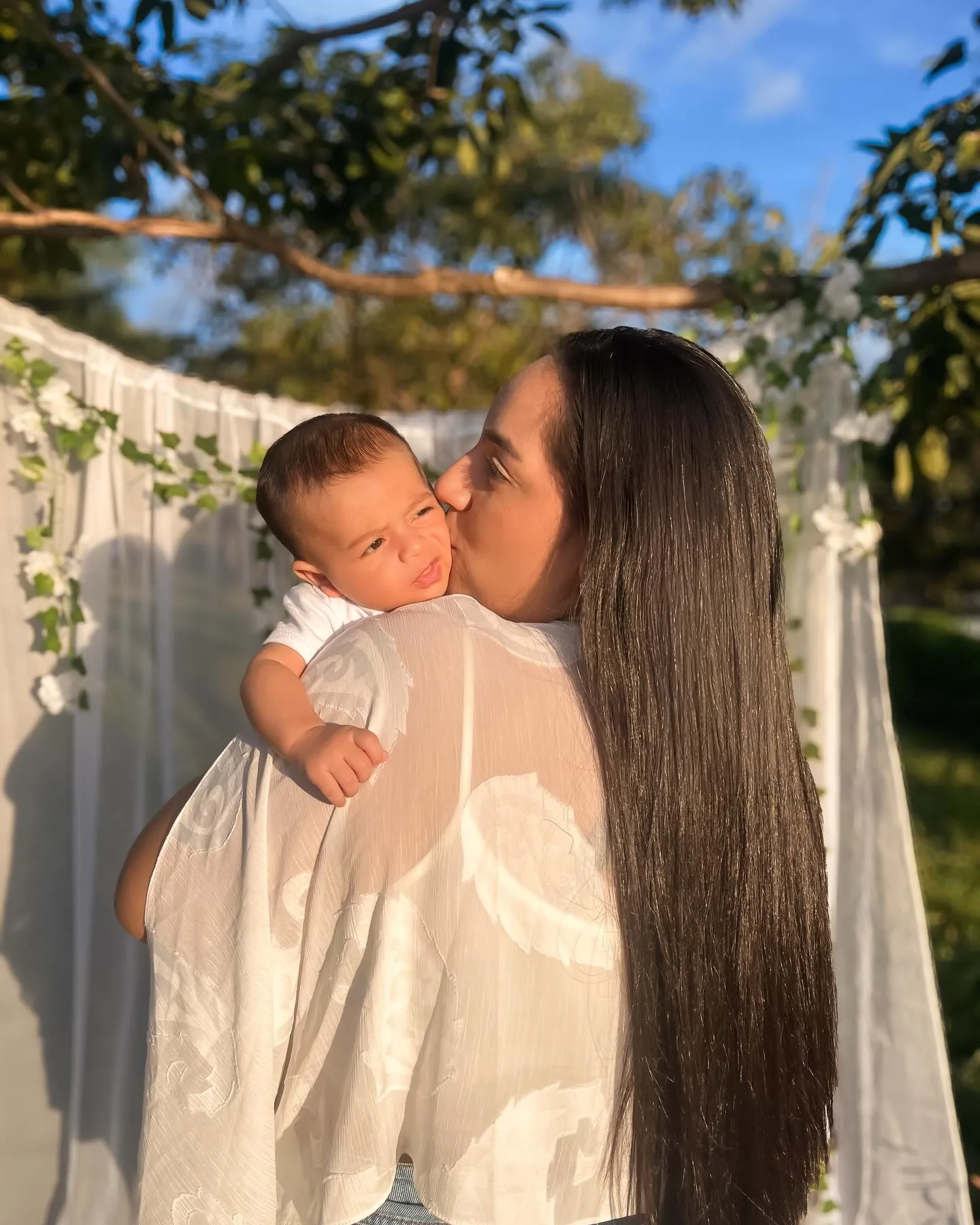 Mulher de roupa branca segura e beija um bebê diante de uma cortina branca ao ar livre.
